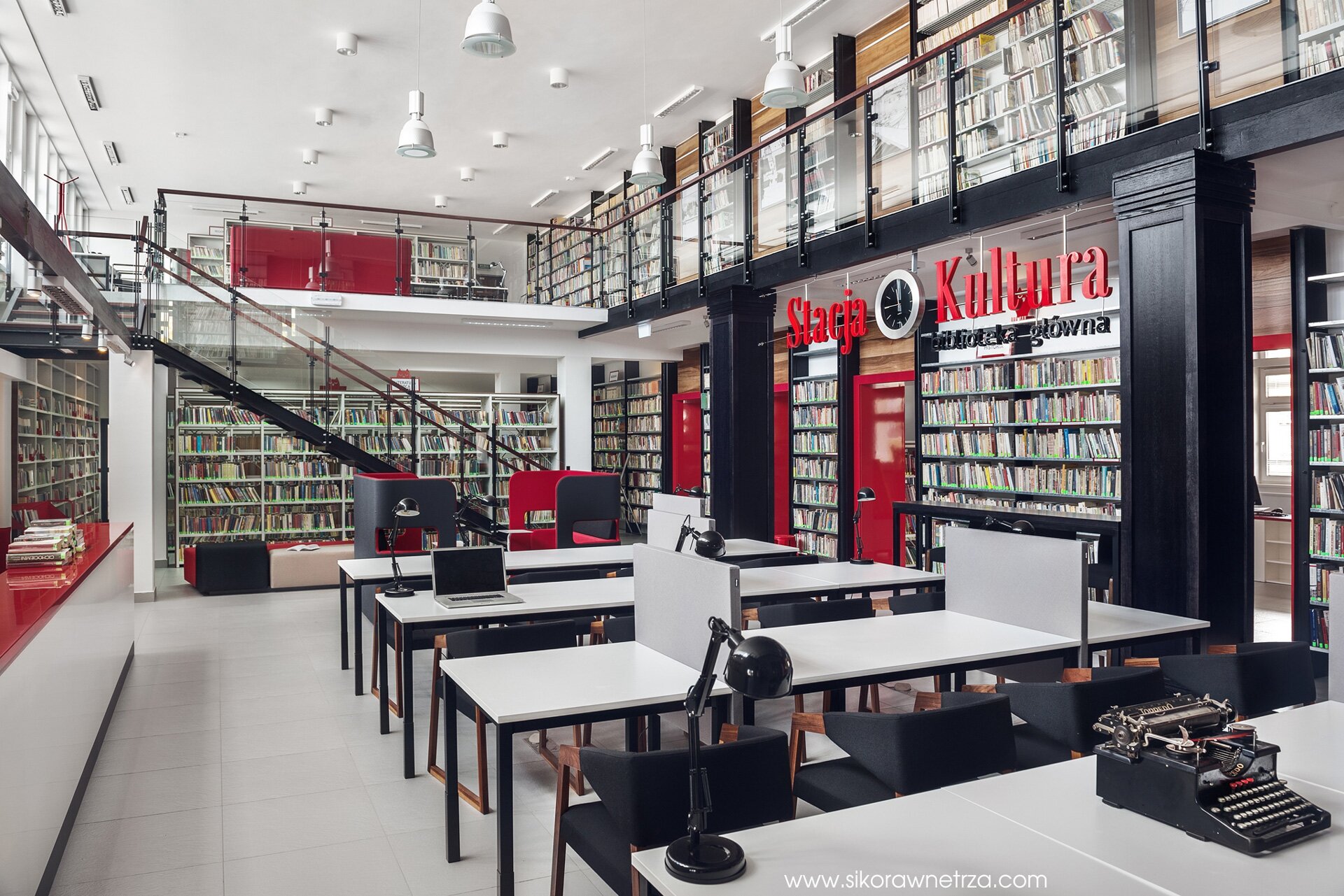 Zdjęcie pomieszczenia w bibliotece. Po bokach ustawione są regały z książkami, na środku stoliki czytelnicze. Po prawej stronie napis Stacja Kultura. Wewnątrz pomieszczenia są schody prowadzące na antresolę, na której stoją regały z książkami.  