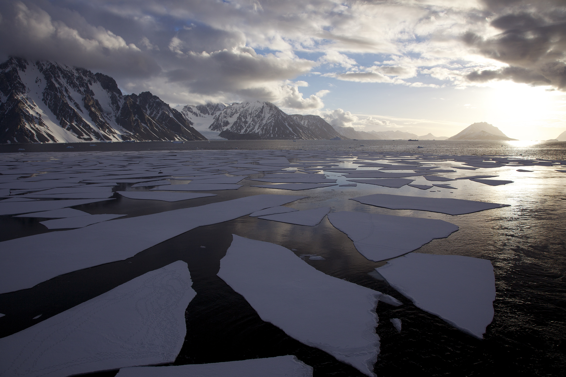 Fotografia prezentuje krajobraz Antarktydy latem. Na pierwszym planie widoczna kra pływająca po morzu. W tle góry pokryte częściowo śniegiem, nad nimi kłębiaste chmury oraz słońce nad horyzontem.