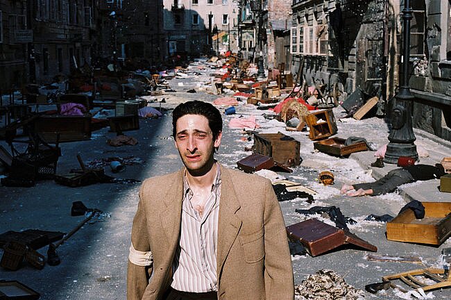 Ilustracja przedstawia idącego opustoszałą ulicą mężczyznę. To młody, szczupły mężczyzna o pociągłej twarzy. Jego oczy są załzawione. Jego włosy są ciemne i zaczesane do tyłu. Mężczyzna ubrany jest w jasną marynarkę i koszulę w paski. Na prawej ręce ma opaskę. Na drugim planie, za mężczyzną znajduje się obraz zniszczonej, zrujnowanej i ograbionej warszawskiej ulicy. Porozrzucane są walizki, meble i ubrania. Pomiędzy nimi leżą ludzkie ciała. W domach przy ulicy pootwierane są okna.