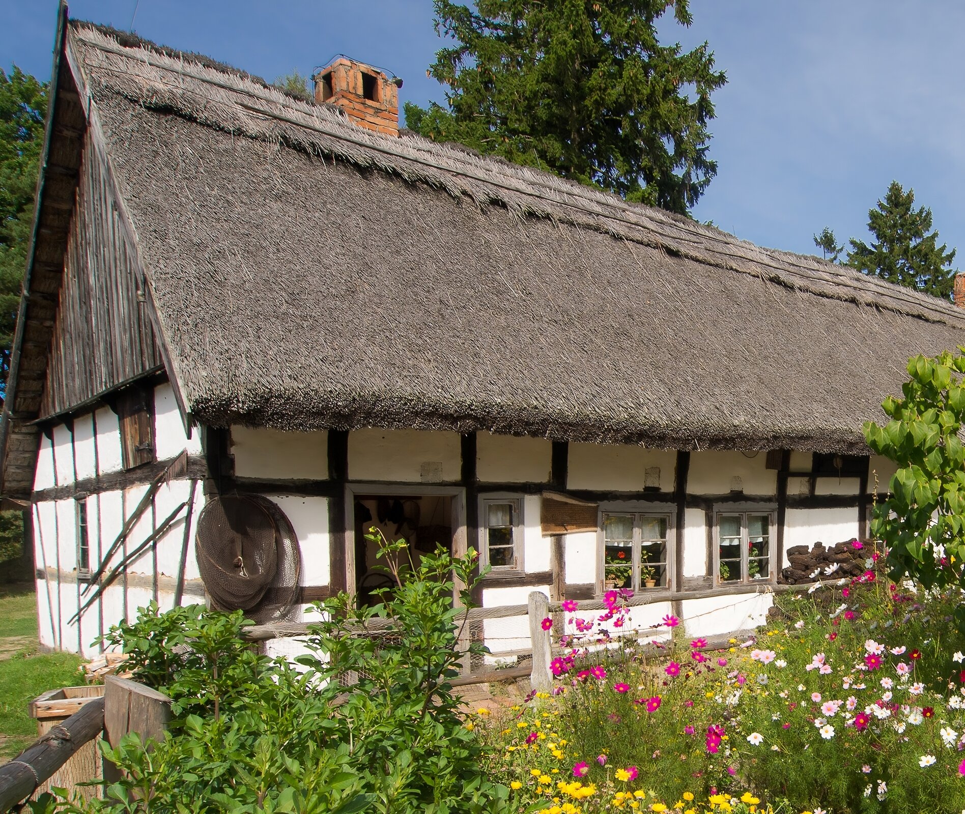 Kliknij, aby powiększyć. Zdjęcie przedstawia fragment chaty. Jest jednopiętrowa i posiada dwuspadowy dach pokryty trzciną. Budynek ma konstrukcję szkieletową złożoną z drewnianych belek wypełnionych murem, który jest bielony. Wokół domu znajduje trawnik, widać też kolorowe kwiaty.