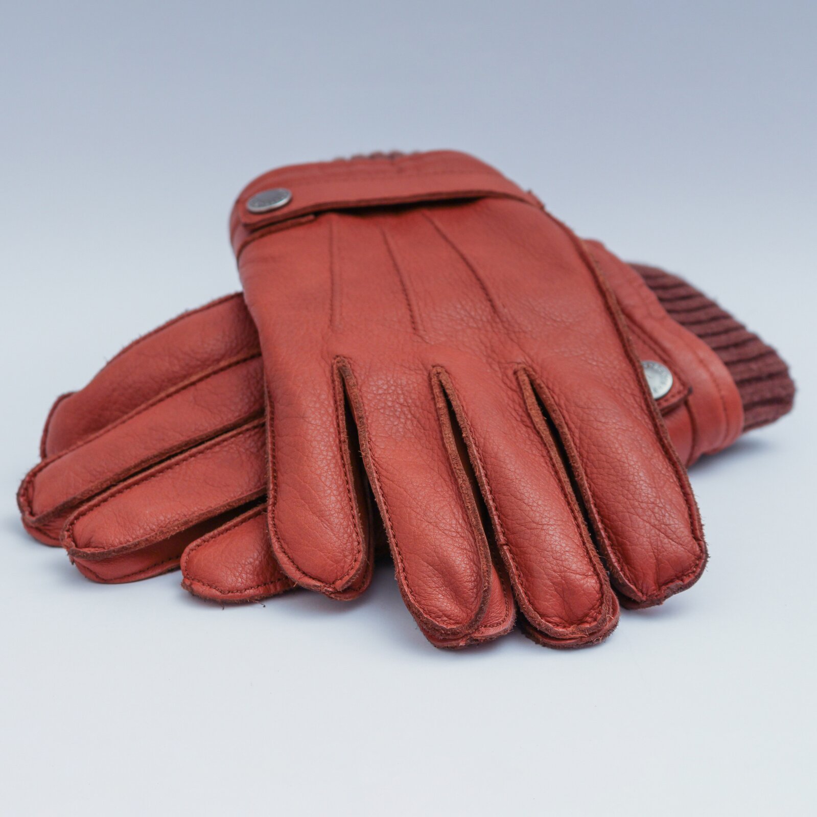 Zdjęcie przedstawia parę skórzanych, zimowych rękawiczek w kolorze brązowym. Rękawiczki mają przy nadgarstku taśmowate zapięcie, zapinane na klips oraz dzianinowy ściągacz.