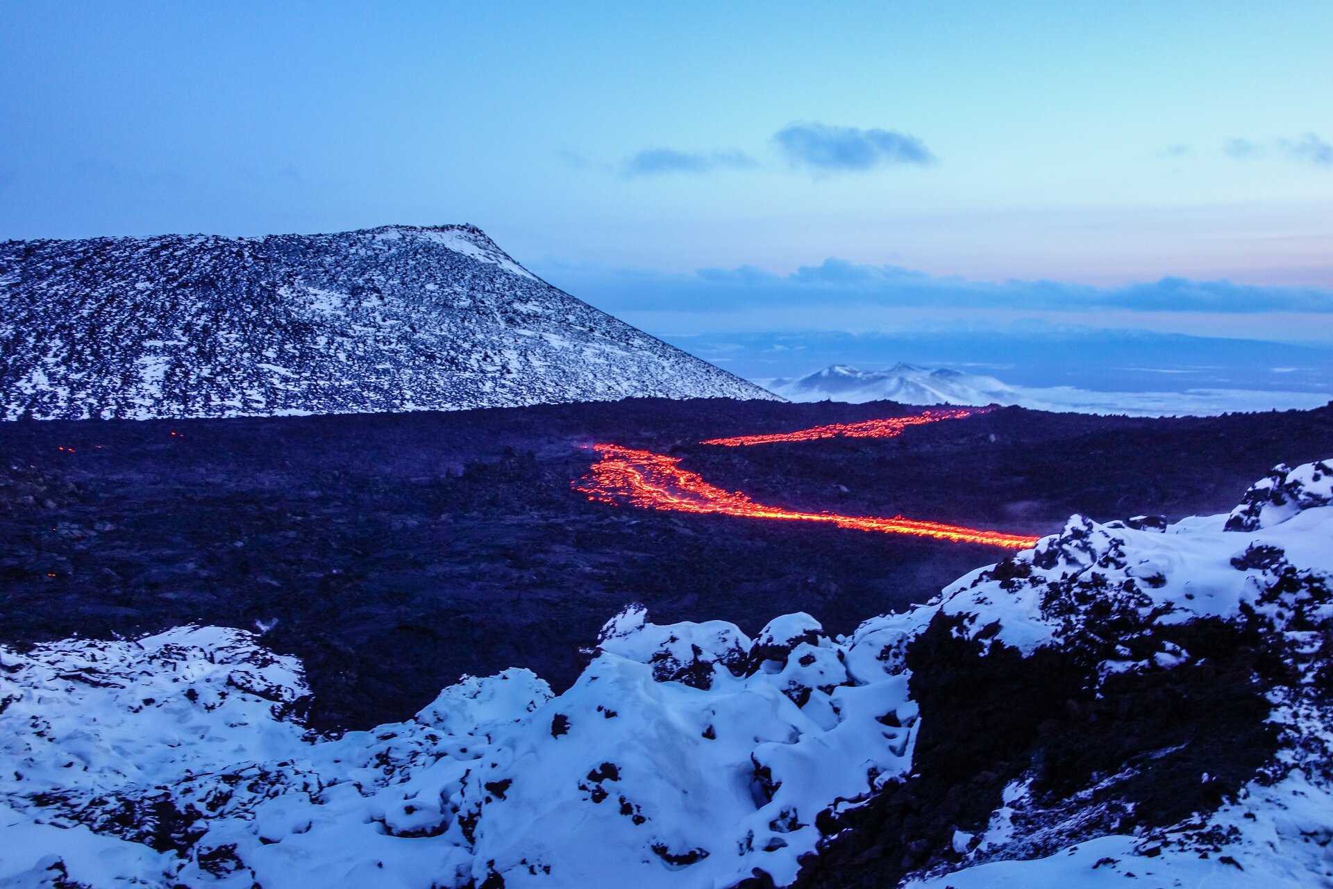 Na fotografii erupcja powierzchniowa na Kamczatce. Szare pole popiołu wulkanicznego otaczają ośnieżone wzniesienia. Wzdłuż płaszczyzny biegnie szczelina, z której wydobywa się płonąca lawa.    