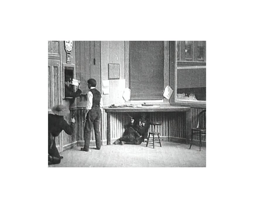 Kadr z czarno-białego filmu. Znajdują się na nim cztery osoby. Po lewej stronie kadru na pierwszym planie widać klęczącego mężczyznę w kapeluszu obróconego tyłem. W dłoni trzyma on pistolet, z którego mierzy do innych uczestników wydarzenia. Na drugim planie po lewej stronie kadru widoczne jest okienko urzędowe, takie jak na poczcie lub w banku. Z okna wygląda mężczyzna w czapce służbowej, w dłoni trzyma białą kartkę lub czek. Przed okienkiem stoi mężczyzna w kamizelce i białej koszuli. Rozmawia z mężczyzną wychylającym się z okienka. Na środku kadru w tle znajduje się stół umieszczony pod oknem. Pod stołem siedzi mężczyzna, który celuje z pistoletu do mężczyzny w kamizelce.  A frame from black and white film. There are four people on it. On the left of the frame in the foreground you can see a kneeling man in a hat turned back. In his hand he holds a pistol, from which he aims at other participants of the event. In the background on the left side of the frame is an official window, such as at the post office or bank. A man in a business cap looks out of the window, holding a white card or check in his hand. A man in a vest and a white shirt is standing in front of the window. He talks to the man leaning out of the window. In the center of the frame in the background is a table placed under the window. Under the table sits a man who is aiming a gun at a man in a vest.