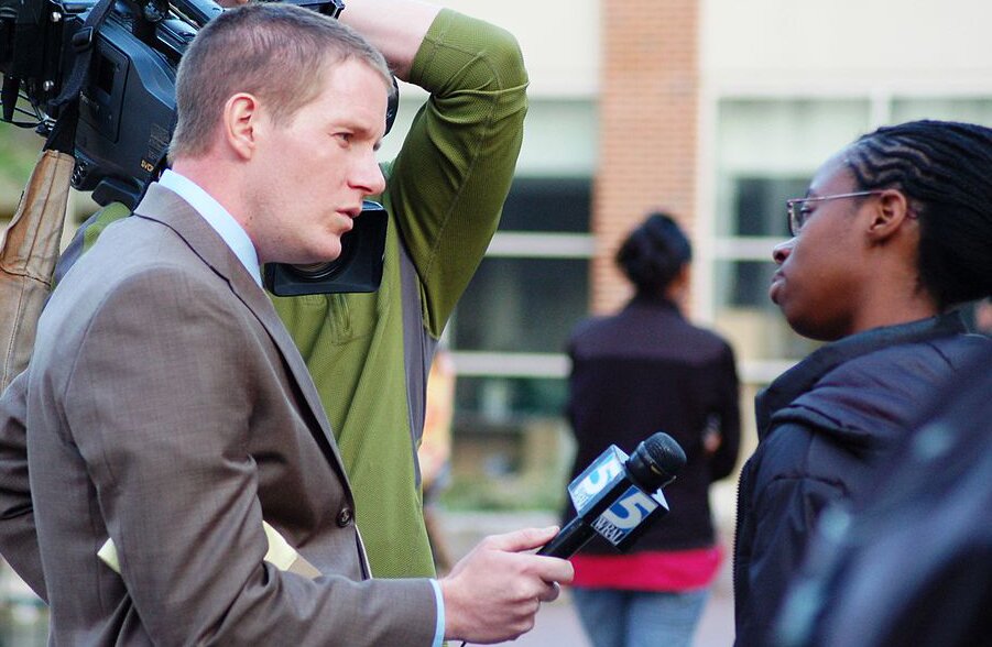 Na zdjęciu młody mężczyzna w marynarce z mikrofonem w ręku. Obok niego stoi mężczyzna operator kamery. Reporter rozmawia z czarnoskórą dziewczyną.  