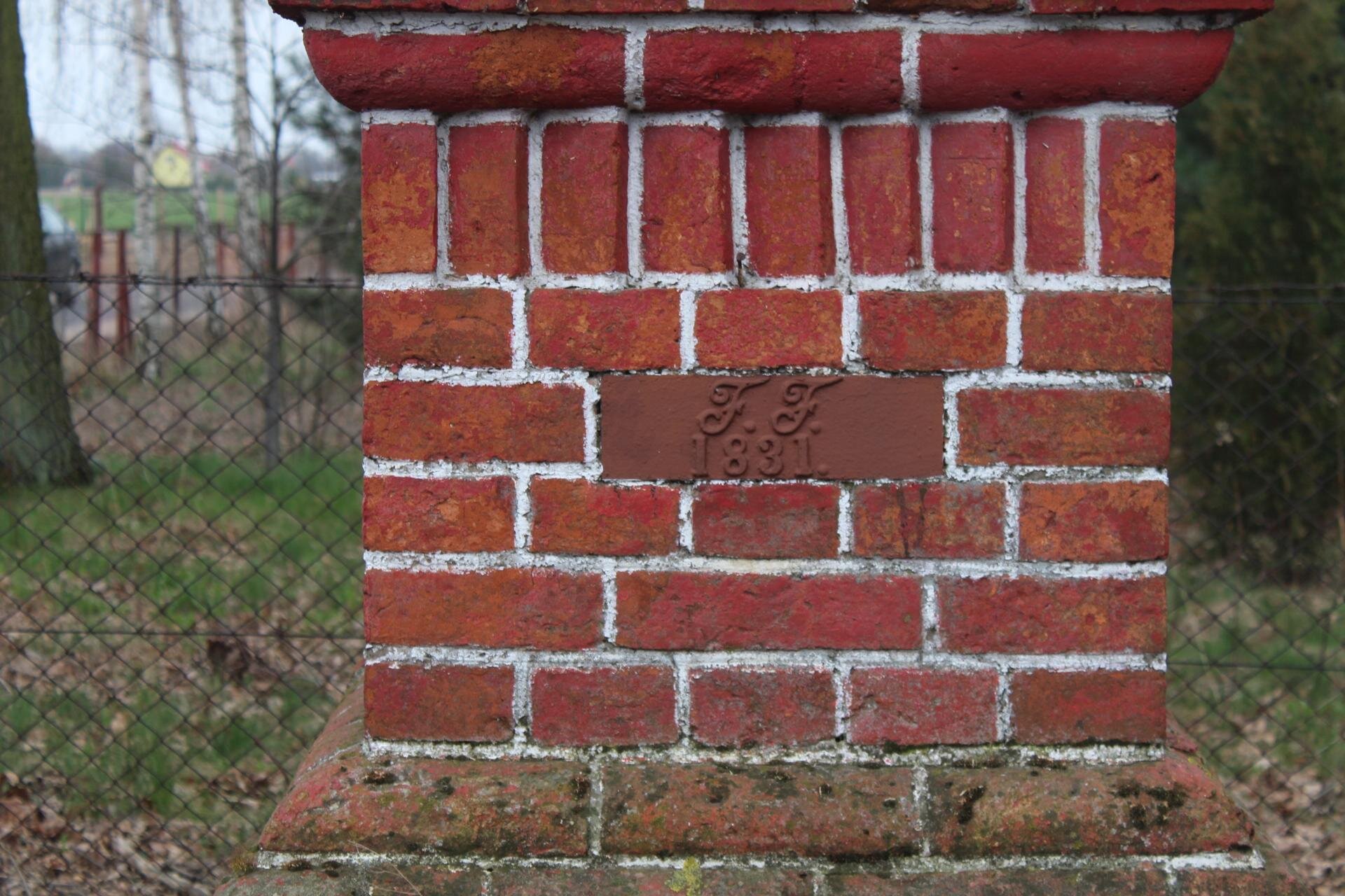 Kolorowe zdjęcie przedstawia postument wymurowany z czerwonej cegły z białymi fugami. W jego centrum widnieje tabliczka z informacją: "F. F. 1831." W tle widać siatkowe ogrodzenie, a za nim trawnik i drzewa.