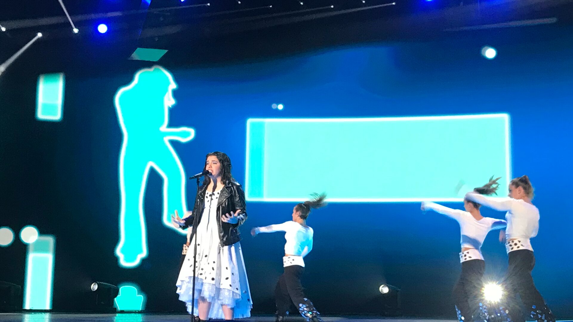 Zdjęcie przedstawia Elisabette Lizza. Jest uśmiechnięta, ma lekko kręcone włosy. Stoi na scenie przed mikrofonem. Ubrana jest w sukienkę z falbaną na dole oraz skórzaną kurtkę. Na scenie za artystką tańczy trzech mężczyzn. Ubrani są w koszule z długim rękawem, długie spodnie i wysokie buty.