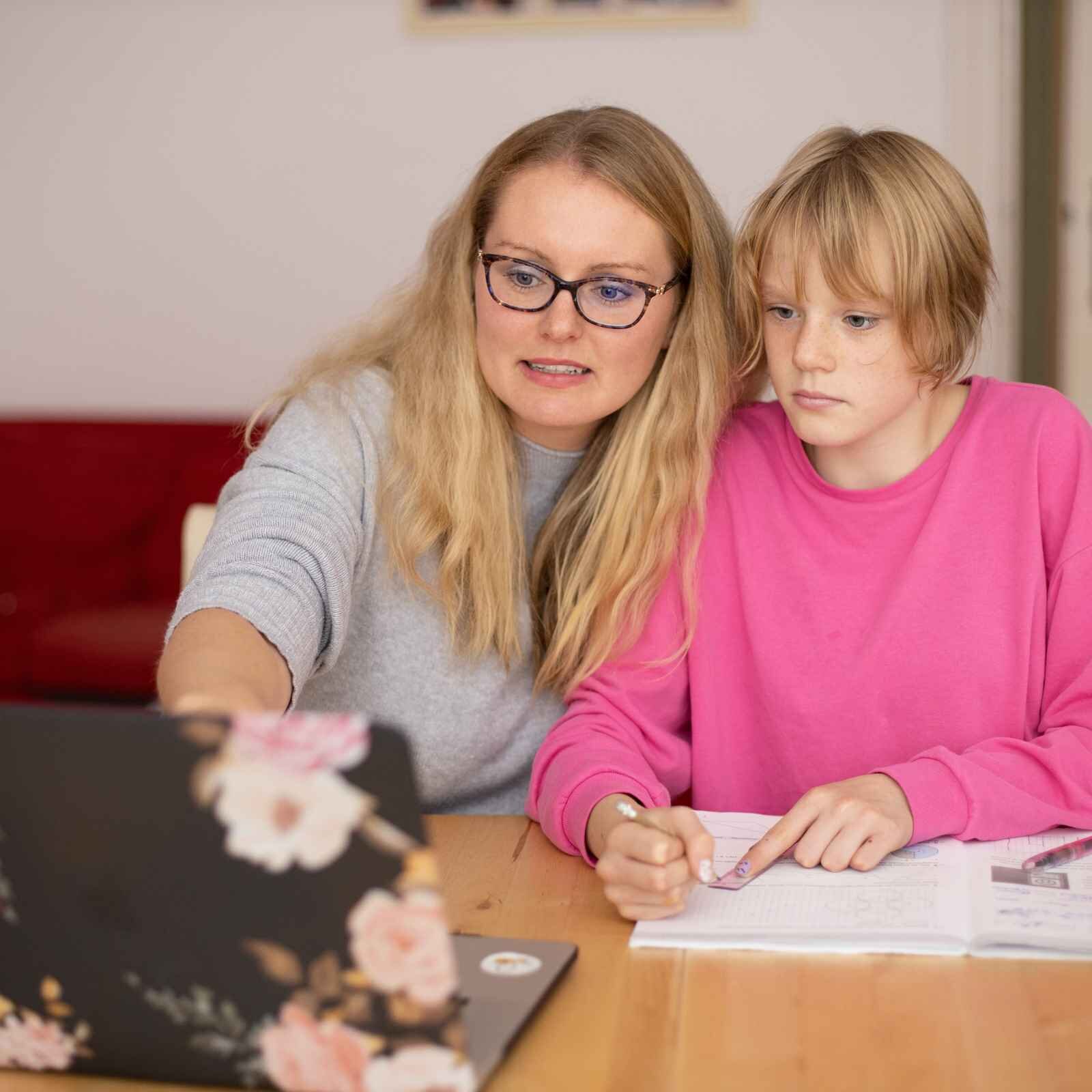 Zdjęcie przedstawia kobietę w średnim wieku oraz nastolatkę, które siedzą przy biurku. Kobieta po lewej stronie ma długie, jasne włosy, okulary korekcyjne i jest ubrana w szary sweterek. Prawą rękę ma wyciągniętą w kierunku stojącego na biurku otwartego laptopa. Nastolatka ma krótkie, jasne włosy i jest ubrana w różową bluzkę. W prawej ręce opartej na blacie trzyma ołówek, a lewą ręką dociska linijkę do zeszytu. Obie spoglądają na monitor laptopa. 