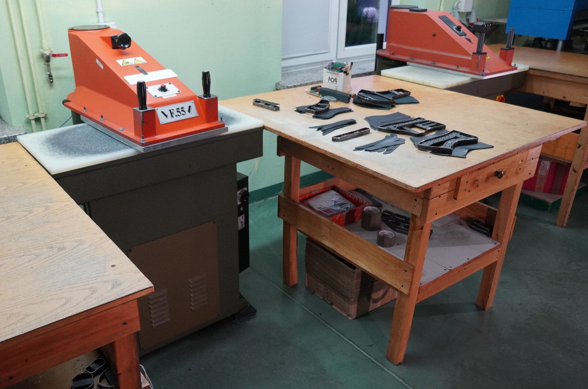 Zdjęcie przedstawia warsztat pracy. Po prawej stronie znajduje się stół, na którym leżą skrawki materiałów, po lewej stronie stoi maszyna. Jest w kolorze pomarańczowym, ma kształt trapezu, z jej przodu znajdują się po obu stronach czarne nieduże bolce. Nagranie tożsame z treścią.