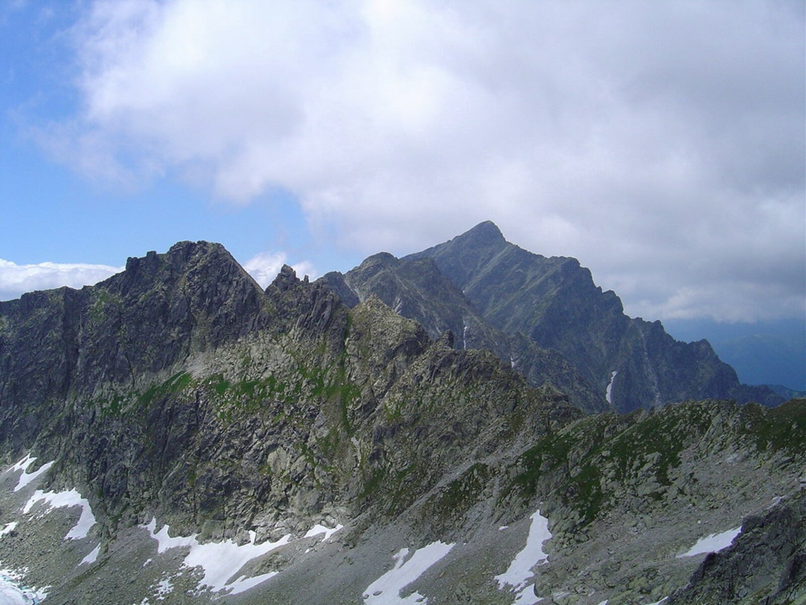 Na zdjęciu tatrzańskie granie. W centralnej części zdjęcia rozległy masyw górski o stromych skalistych zboczach. Skały spękane, o chropowatej strukturze, gdzieniegdzie pokryte niewielką ilością śniegu, a także zielonymi porostami. Granie o charakterystycznych strzelistych, ostrych kształtach.