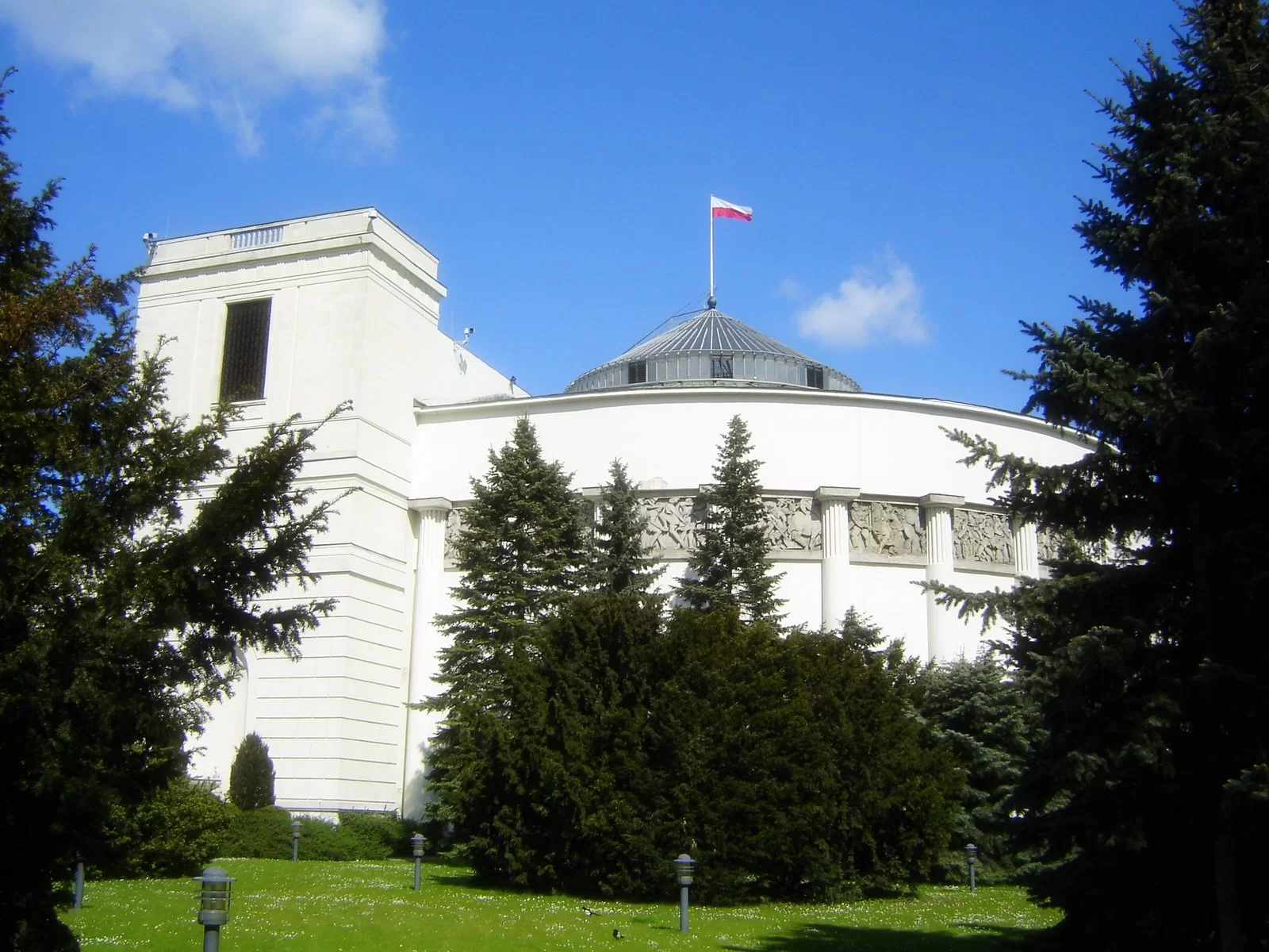 Zdjęcie przedstawia gmach polskiego Sejmu. Widoczna jest duża rotunda, w której mieści się sala obrad. Na szczycie rotundy powiewa polska flaga.