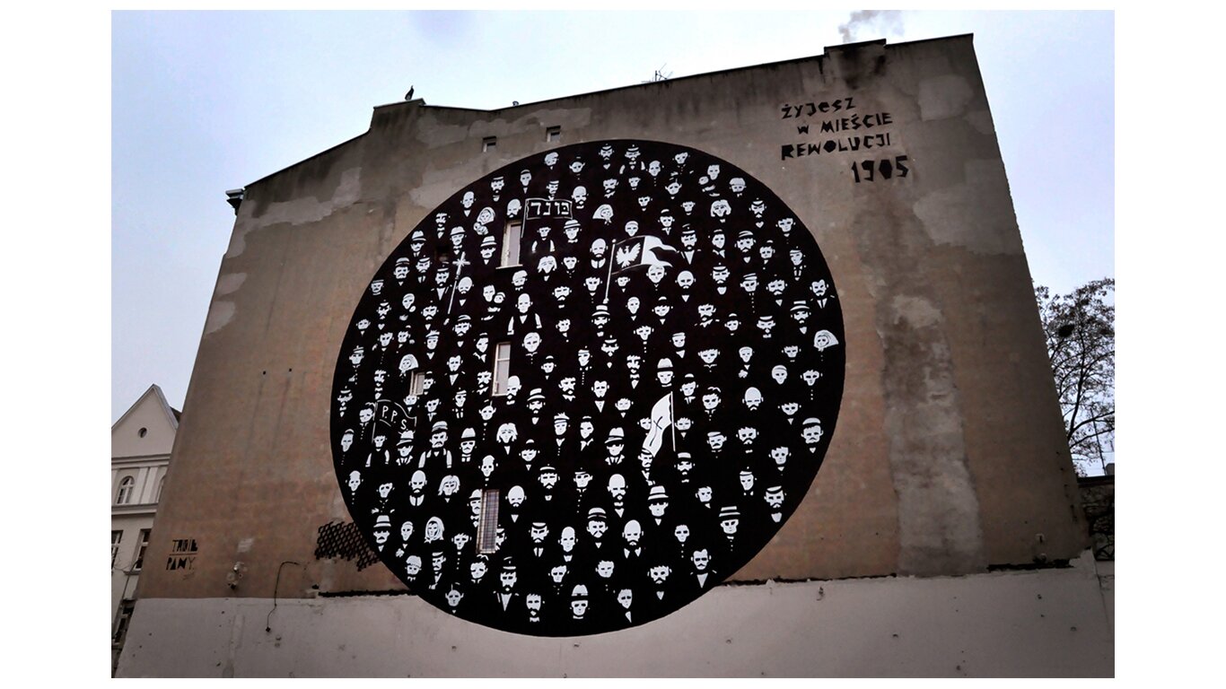 Fotografia barwna przedstawiająca mural  w Łodzi poświęcony rewolucji 1905 roku. Czarne koło, w którym umieszczone są białe twarze ludzi oraz flagi z godłem Polski, napisami w jidysz oraz z napisem PPS. Obok napis: Żyjesz w mieście rewolucji 1905.