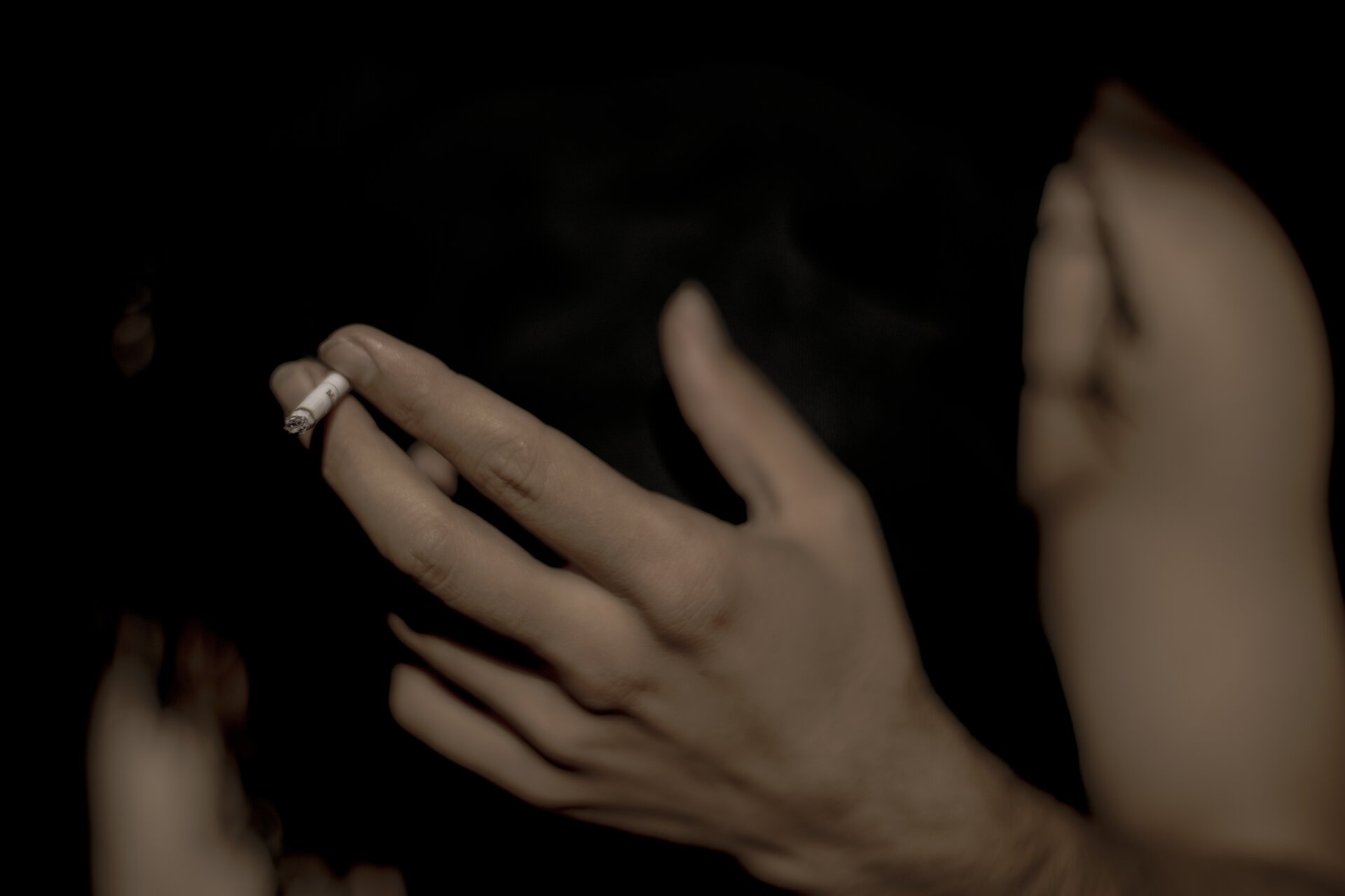 Na zdjęciu widać dłoń trzymającą tlącego się papierosa. Tło zdjęcia jest przyciemnione.