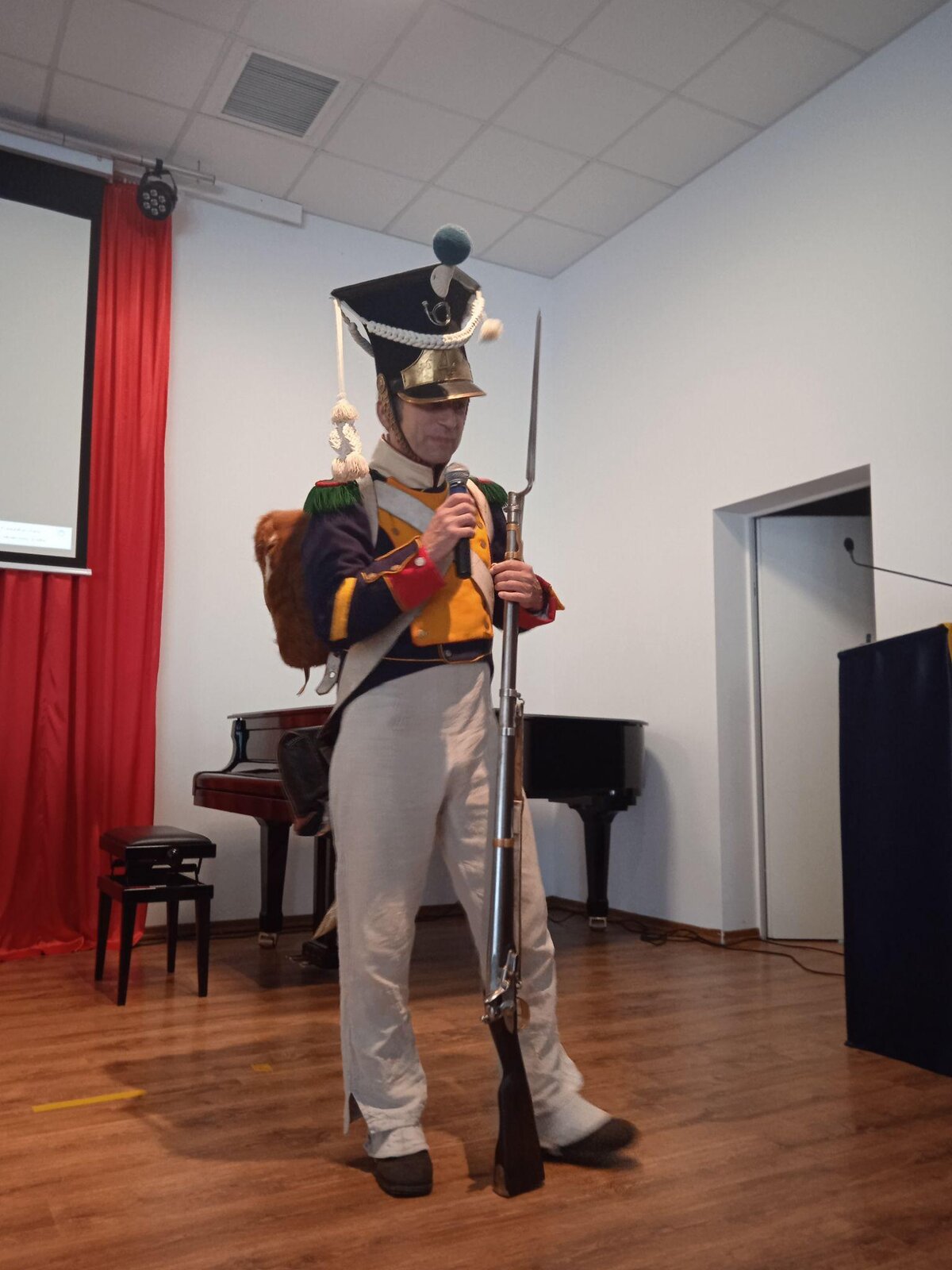 Zdjęcie przedstawia mężczyznę ubranego w stary mundur żołnierski. Mężczyzna ma białe, długie spodnie częściowo zakrywające czarne buty. Ma na sobie granatowo‑żółtą marynarkę, a na plecach brązowy plecak. Na głowie ma wysoką, granatowo‑biało‑złotą czapkę. W lewej ręce trzyma broń z bagnetem na lufie. W prawej dłoni trzyma mikrofon. W tle widoczna jest biała ściana, przy której stoi pianino oraz taboret. Na ścianie widoczny jest fragment czerwonego materiały, na którym znajduje się ekran do wyświetlania zdjęć na rzutniku.