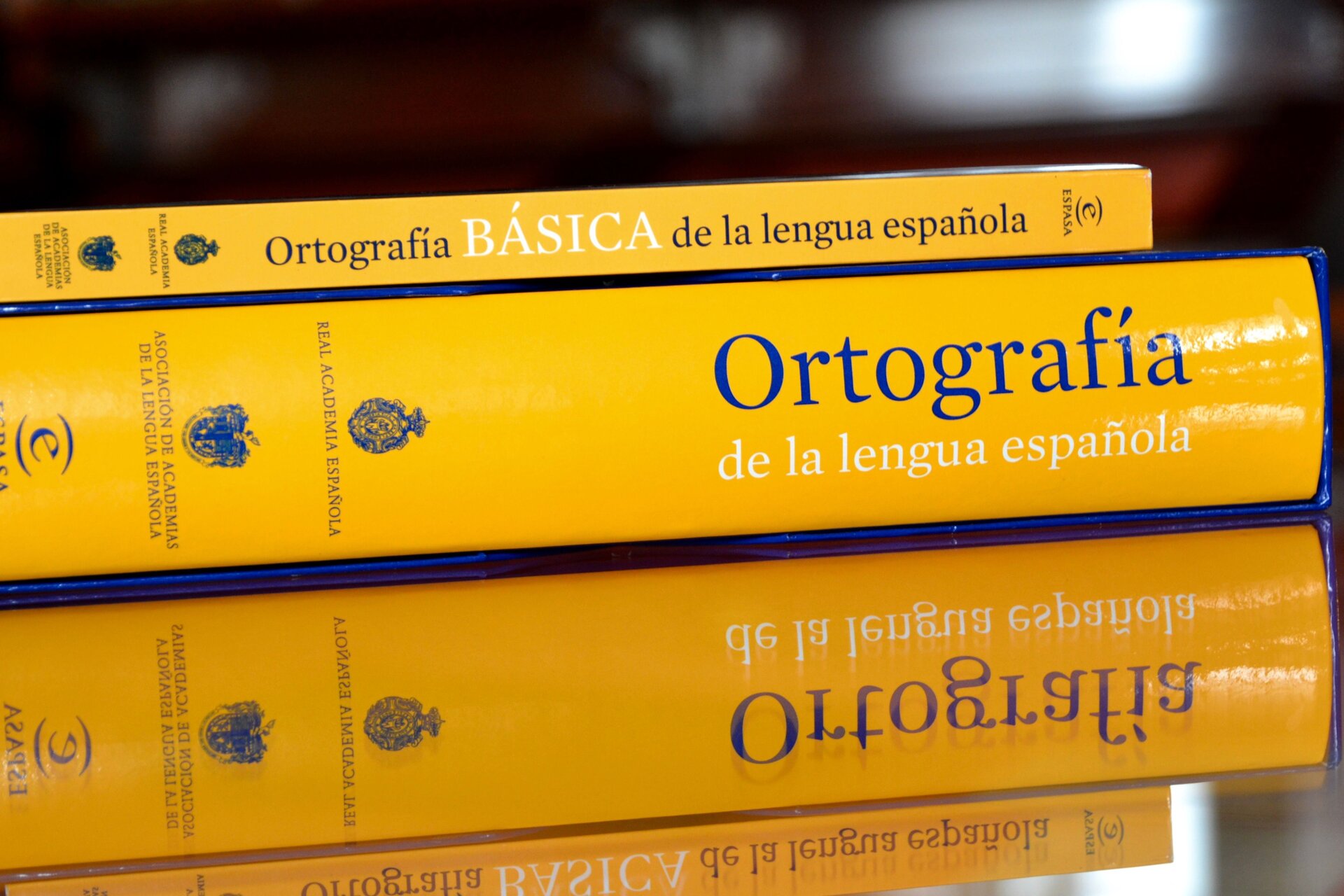 Zdjęcie przedstawia grzbiety dwóch książek. Ich okładki są w żółtym kolorze. Książki są w języku hiszpańskim. Leżą na stole, którego blat jest lustrzany. Daje to złudzenie, jakby na stole znajdowały się nie dwie, a cztery książki. 
