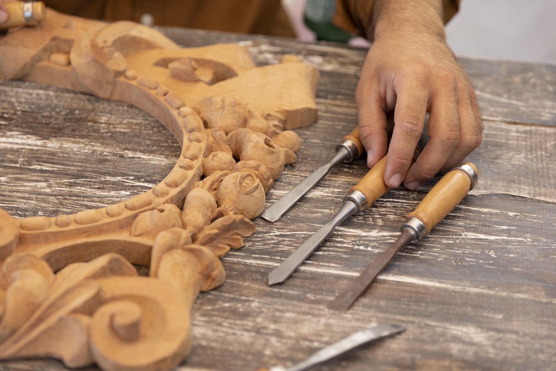 Zdjęcie przedstawia model detalu architektonicznego wykonany z drewna. Model ma kształt półokręgu i wypełniony jest zdobnymi motywami florystycznymi. Na zdjęciu widoczna jest ręka pracownika, która sięga po jedno z dłut położonych na stole obok modelu.