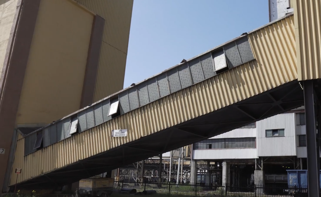 Na zdjęciu widać miejsce połączenia zbiornika magazynowego z łącznikiem. Zbiornik jest wielkokubaturowym obiektem o kształcie prostopadłościanu. W dolnej części obiektu z budynkiem łączy się pochylnia. Pochylnia jest o kształcie prostopadłościanu zapadającego w stronę gruntu. W tle widać obiekty przemysłowe.