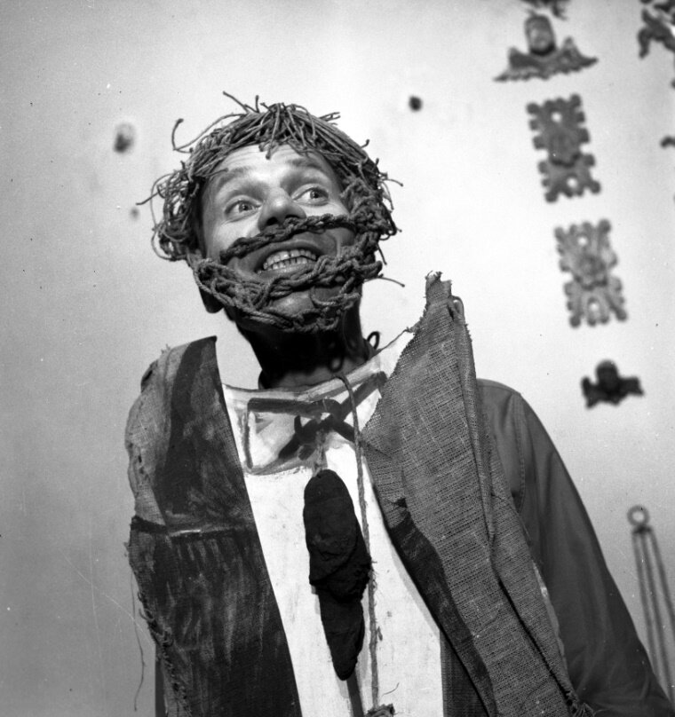 Zdjęcie przedstawia mężczyznę. To aktor wcielający się w rolę Poety w Weselu Wyspiańskiego. Mężczyzna ma szeroko otwarte oczy. Szczerzy zęby. Na głowie ma perukę zrobioną z włóczki. Również jego broda i wąsy zrobione są z włóczki. Mężczyzna stoi przed wieszakiem, na którym zawieszony jest niby-garnitur. Jest on zrobiony ze strzępów materiału. Jest cały poszarpany.