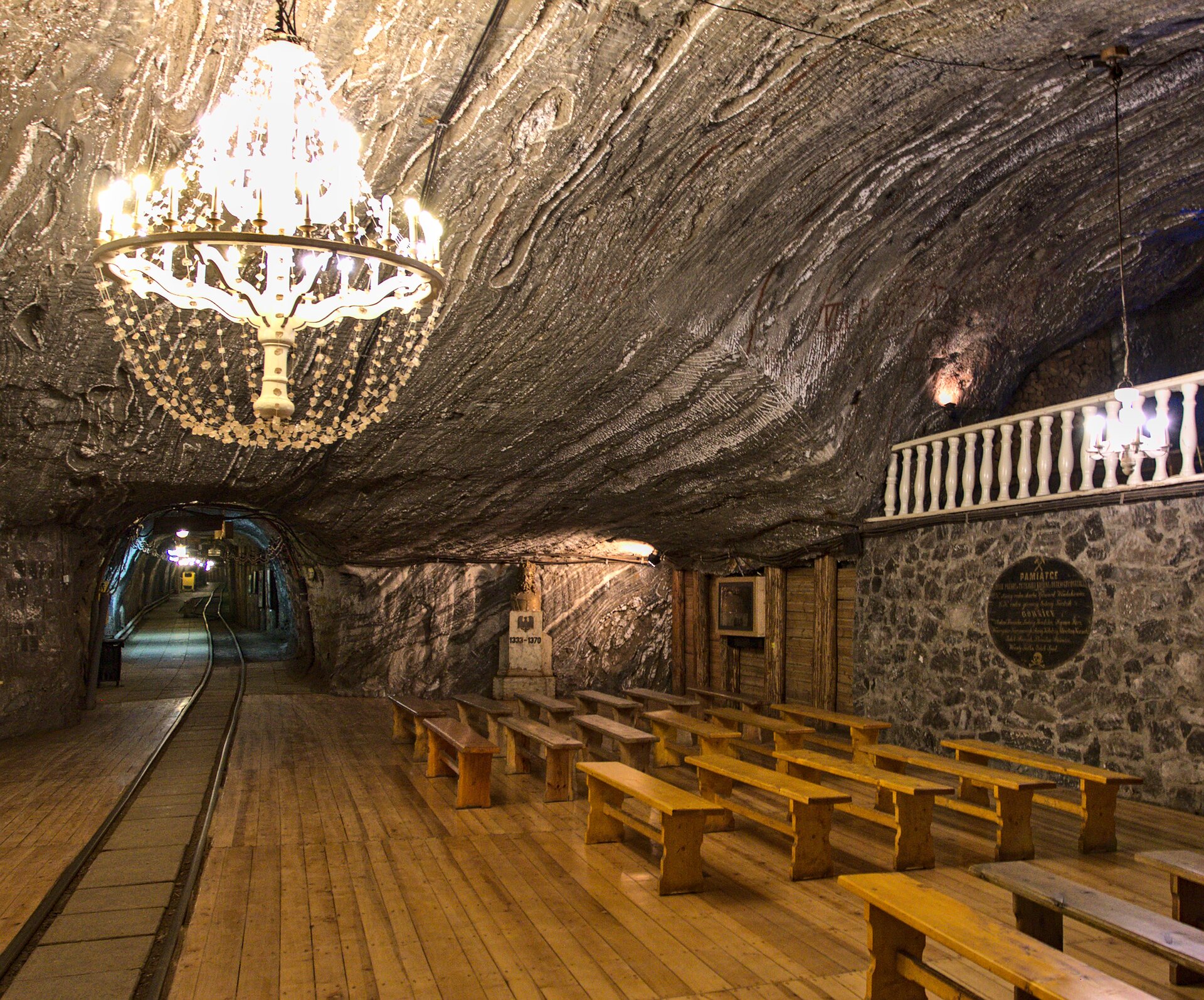 Zdjęcie przedstawia część Kaplicy Świętej Kingi. Strop jest stosunkowo niski, skalny. Po prawej stronie zdjęcia stoją kościelne ławki, po lewej stronie zdjęcia są wąskie tory kolejowe. Ze stropu zwisa żyrandol.