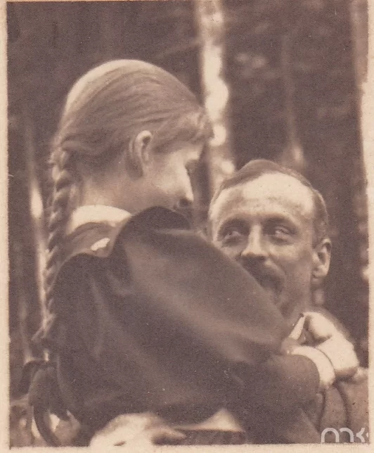 Fotografia przedstawia mężczyznę w sile wieku trzymającego w ramionach dziewczynkę. Postać ukazuje lewy połprofil owalnej twarzy. Włosy mężczyzny są ciemne, proste, zaczesane na prawą stronę. Ma on lekko zmarszczone czoło, jasne brwi i oczy zwężone w uśmiechu. Patrzy na córkę. Nos mężczyzn jest prosty. Pisarz ma wąsy. Mężczyzna jest ubrany w marynarkę. Dziewczynka ukazuje prawy profil. Ma lekko opuszczone powieki i niewielki nos. Jej włosy są długie, zaplecione w warkocz. Dziecko obejmuje oburącz szyję mężczyzny. Dziewczynka jest ubrana w sukienkę. W tle ukazane są wysokie drzewa.