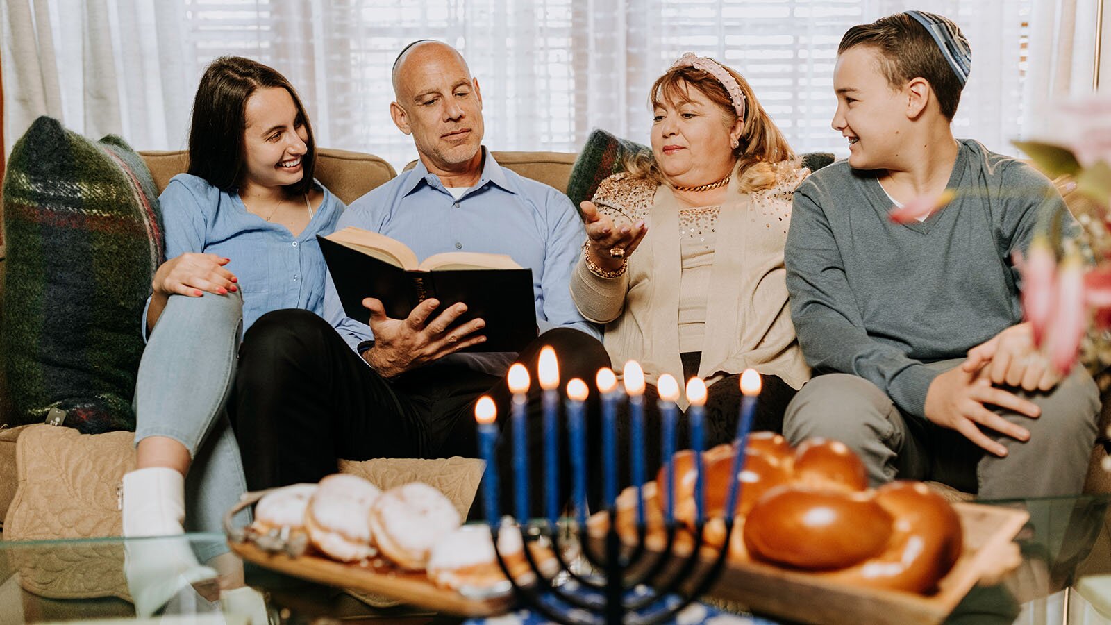 Zdjęcie przedstawia rodzinę cztery osoby siedzącą wspólnie na kanapie. Od prawej widoczna jest młoda dziewczyna, starszy mężczyzna trzymający w ręku książkę, starsza kobieta z podniesioną do góry prawą dłonią oraz młody chłopak, zwrócony w ich kierunku, z jarmułką na głowie. Przed nimi widoczny jest niski stół, na którym widoczny jest świecznik dziewięcioramienny z palącymi się cienkimi, niebieskimi świeczkami, chałka w drewnianej podłużnej misce oraz pączki posypane cukrem pudrem ułożone na drewnianej podstawce.
