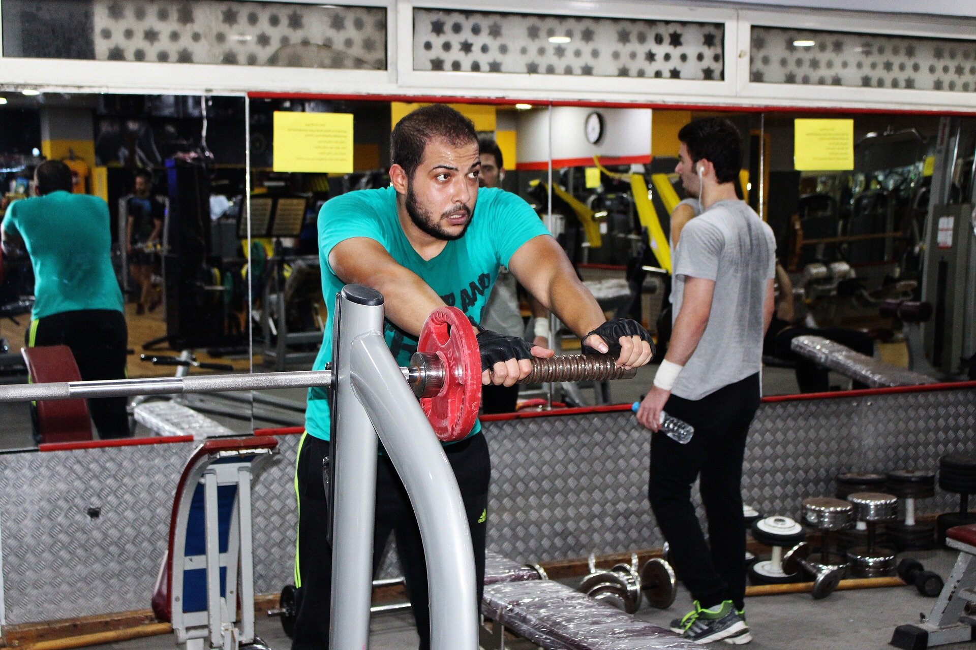 Zdjęcie przedstawia młodego mężczyznę znajdującego się na sali ćwiczeń w siłowni. Mężczyzna ubrany jest w sportowy strój, ma lekką nadwagę. Jest spocony, jego twarz wyraża niezadowolenie, zmęczenie i nadmierny wysiłek. 