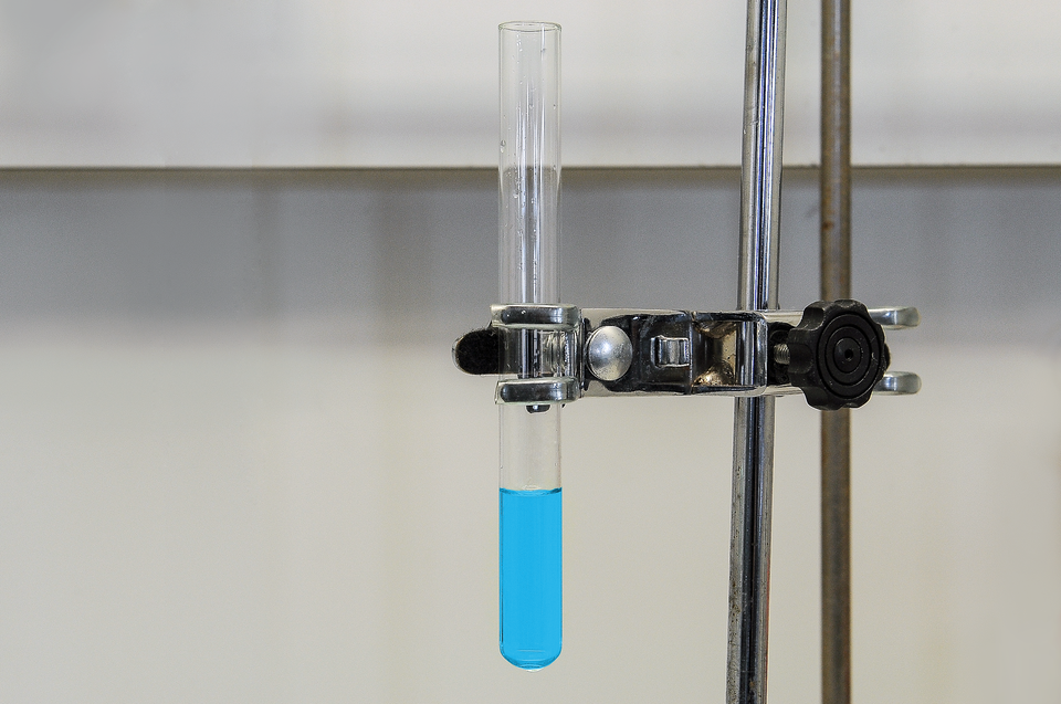 Grafika przedstawia zdjęcie wykonane w laboratorium. Na zdjęciu widoczna jest szklana bezbarwna probówka zawieszona metalową łapą  na metalowym statywie. Wewnątrz probówki, do jednej trzeciej jej objętości, znajduje się niebieska ciecz.