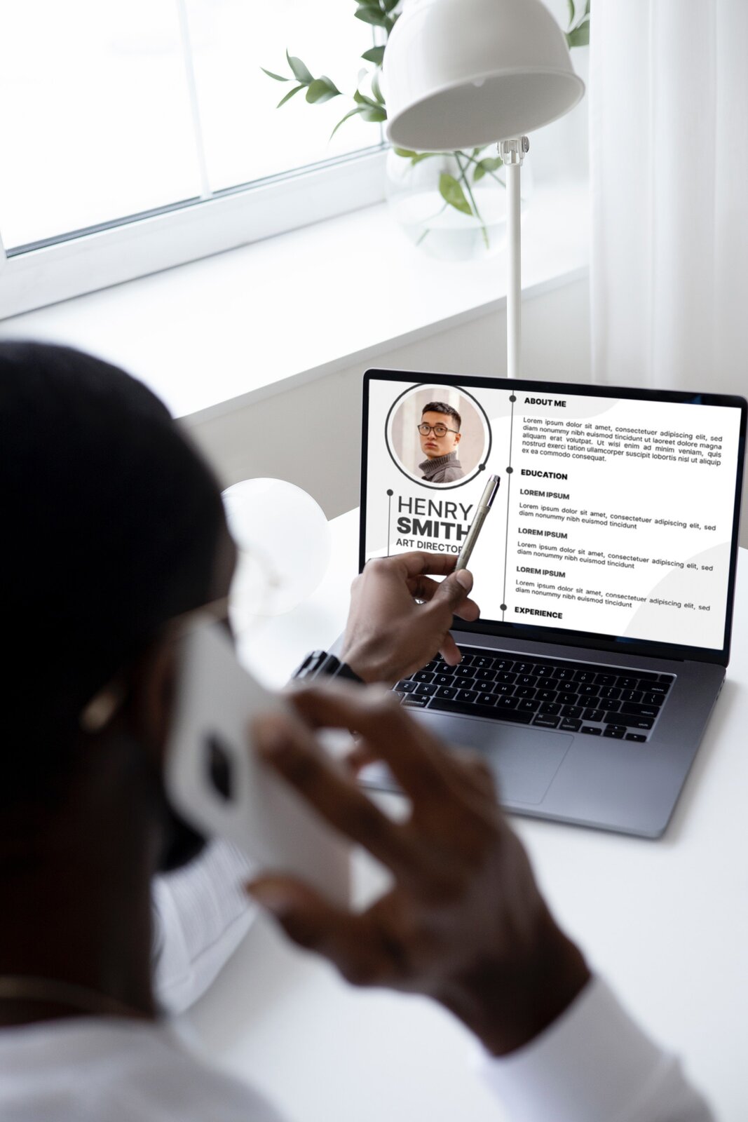 Zdjecie przedstawia rekrutera rozmawiającego przez telefon. Siedzi przy biurku i ma włączony komputer przenośny . Na ekranie wyświetla się cv należące do HENREGO SMITH.  Widoczne są napisy w języku angielskim ABOUT ME, EDUCATION, EXPERIENCE oraz zdjęcie twarzy młodego mężczyzny. 