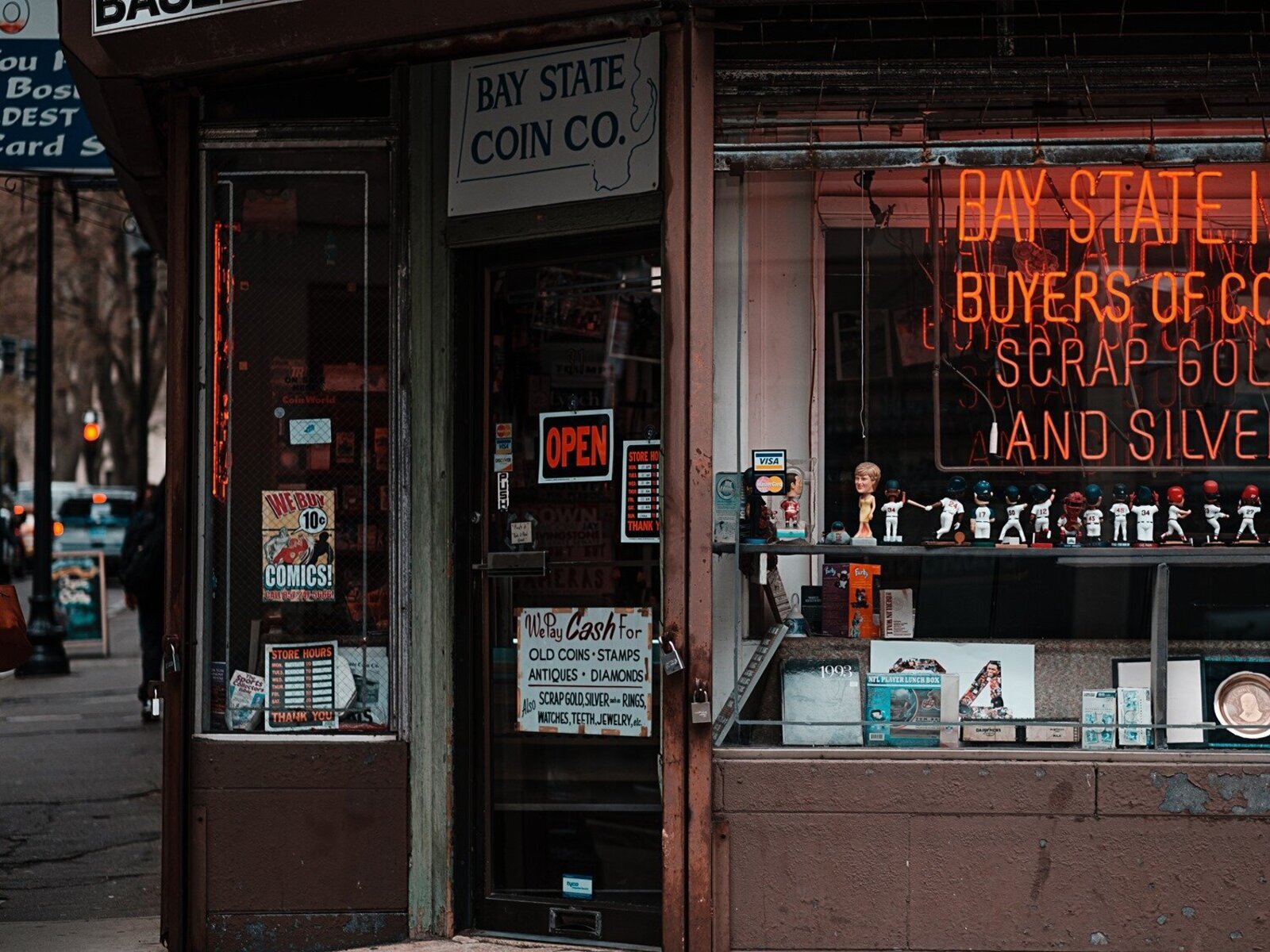 Zdjęcie przedstawia starą witrynę narożnego sklepu ze starociami oraz wejście do tego sklepu. W witrynie ustawione są niewielkie figurki sportowców, pudełka z grami, komiksy. Na wejściu widoczna jest tabliczka z napisem w języku angielskim: OPEN, a poniżej szyld z napisem w języku angielskim: We Pay Cash for OLD COINS, STAMPS, ANTIQUES, DIAMONDS. Also SCRAP GOLD, SILVER such as RINGS, WATCHES, TEETH, JEWELRY, etc. Na wejściem wisi szyld z napisem w języku angielskim: BAY STATE COIN CO. Po prawej stronie od wejścia w oknie wisi komiksowy plakat z napisem w języku angielskim:WE BUY COMICS.