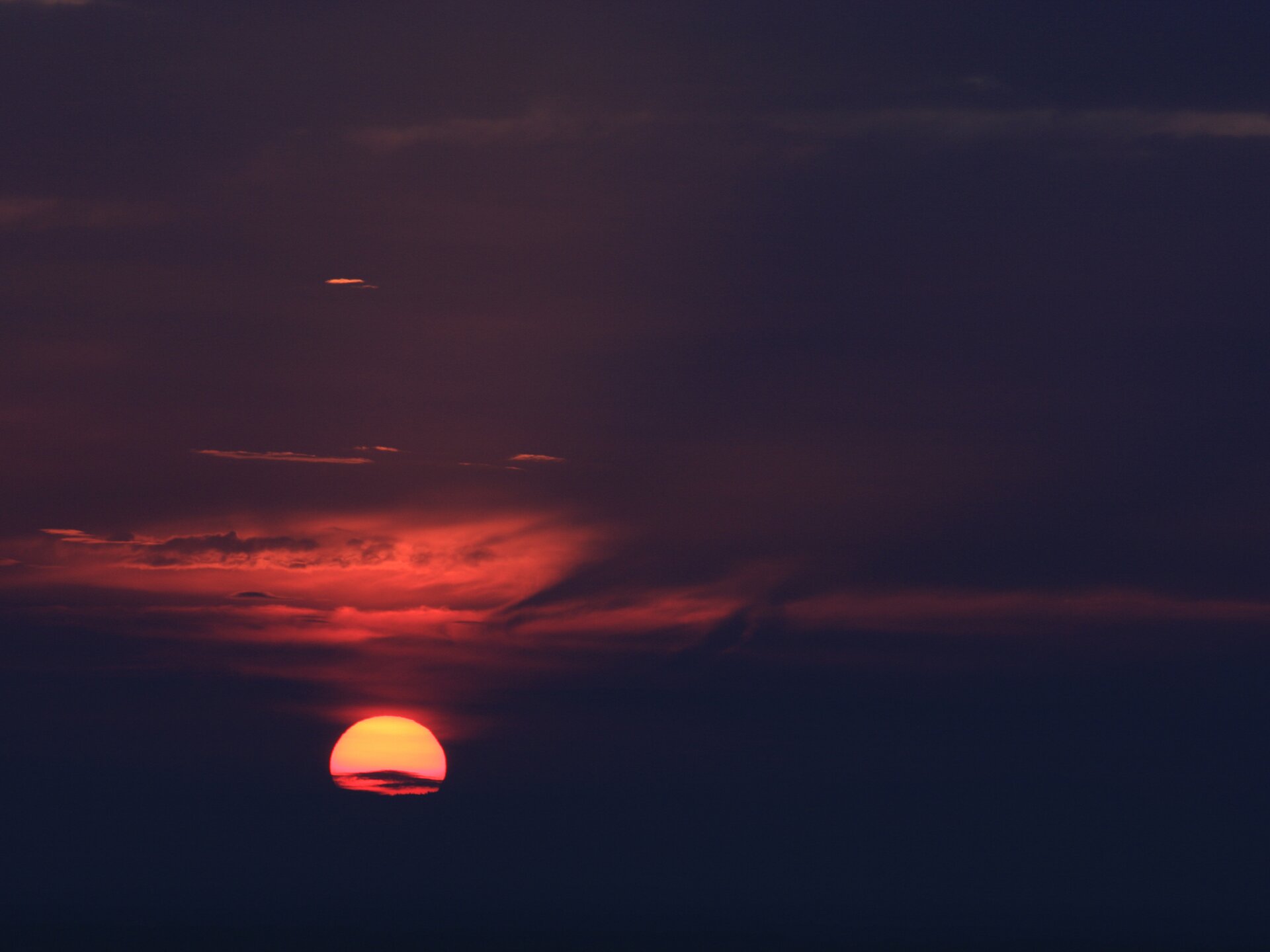 Zdjęcie przedstawia niebo, a na nim zachodzące słońce. Dolnym skrajem schowało się już za chmurami na horyzoncie. Znajduje się na środku dolnej lewej ćwiartki kompozycji. Chmury ponad nim przybrały barwę głębokiej czerwieni do połowy wysokości kadru. Wyżej, jego lewą stronę rozjaśnia lekka poświata. W niej pojedyncze jasne wąskie pasemko obłoku. Reszta fotografii tonie w mroku. Widok jest nienaturalnie ciemny.