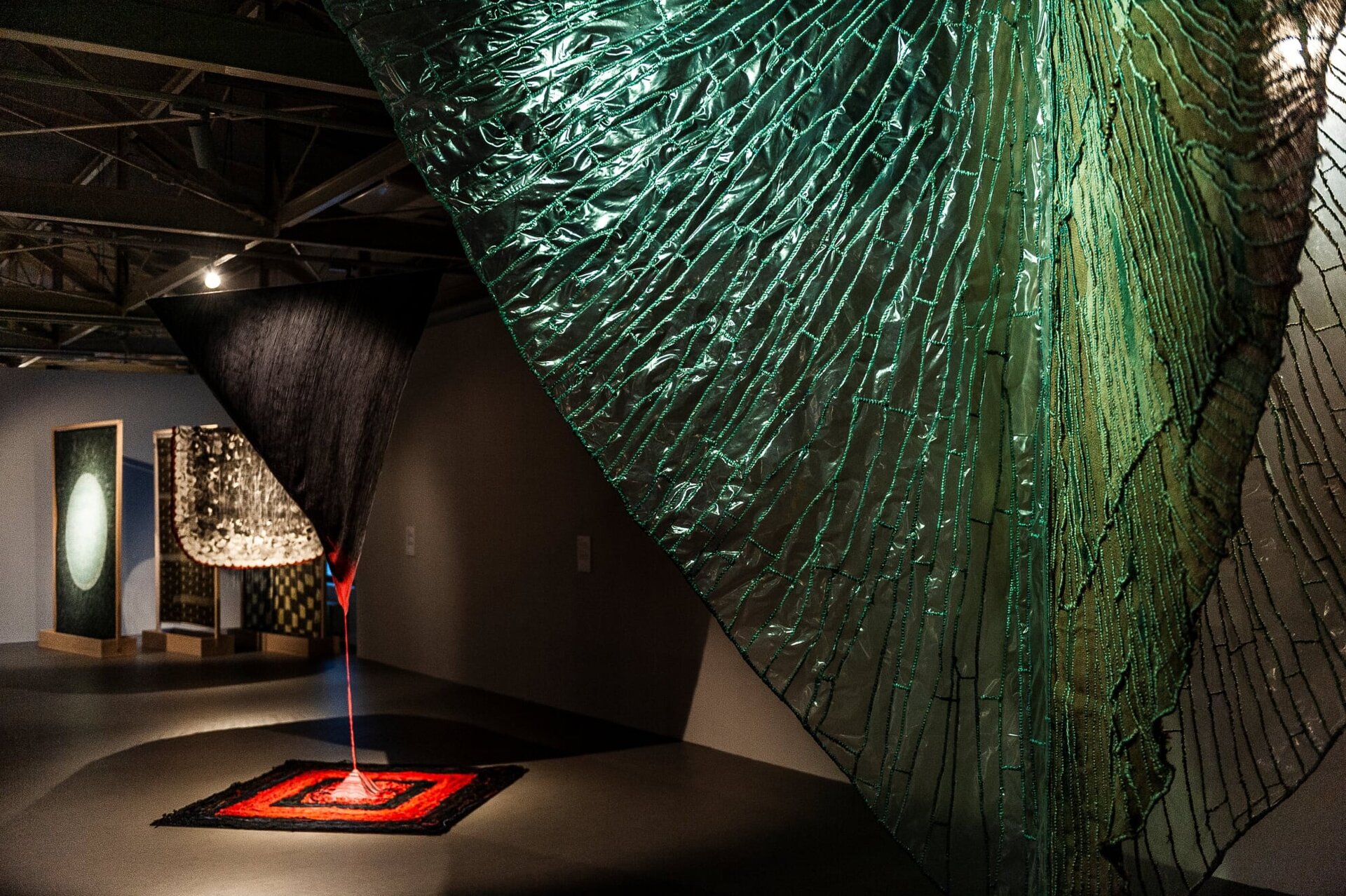 Na zdjęciu ukazano salę wystawienniczą. Na pierwszym planie znajduje się fragment tkaniny w kolorze zielonym. Jest on delikatnie prześwitujący z wieloma przeszyciami zieloną nitką. Przeszycia te tworzą nieregularne czworoboki. Na drugim planie znajduje się wiszący materiał w kształcie trójkąta. Jest on prawie cały w kolorze czarnym. Jedynie dolny róg jest w kolorze czerwonym. Od tego miejsca odchodzi pionowa, czerwona linia. Łączy się z materiałem, który jest poniżej. Ma on kształt kwadratu i jest czarno‑czerwony. W tle znajdują się ciemne, wzorzyste materiały w drewnianych ramach. Tkaniny znajdują się w ciemnym pomieszczeniu.