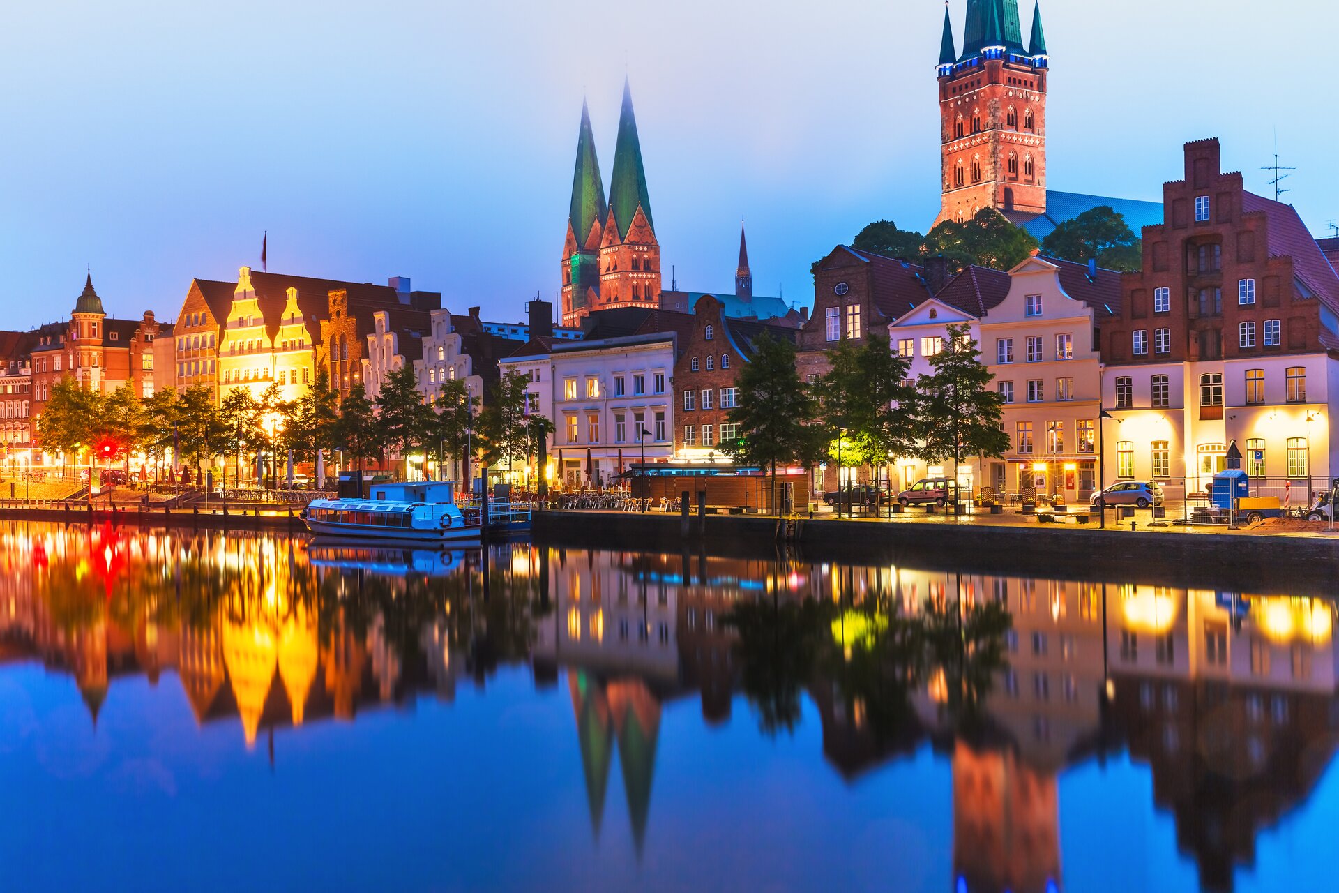 Zdjęcie przedstawia widok na miasto Lubecka, które znajduje się w Niemczech. Zdjęcie wykonano w nocy. Budynki są pieknie podświetlone i ich kształt odbija się w wodzie.