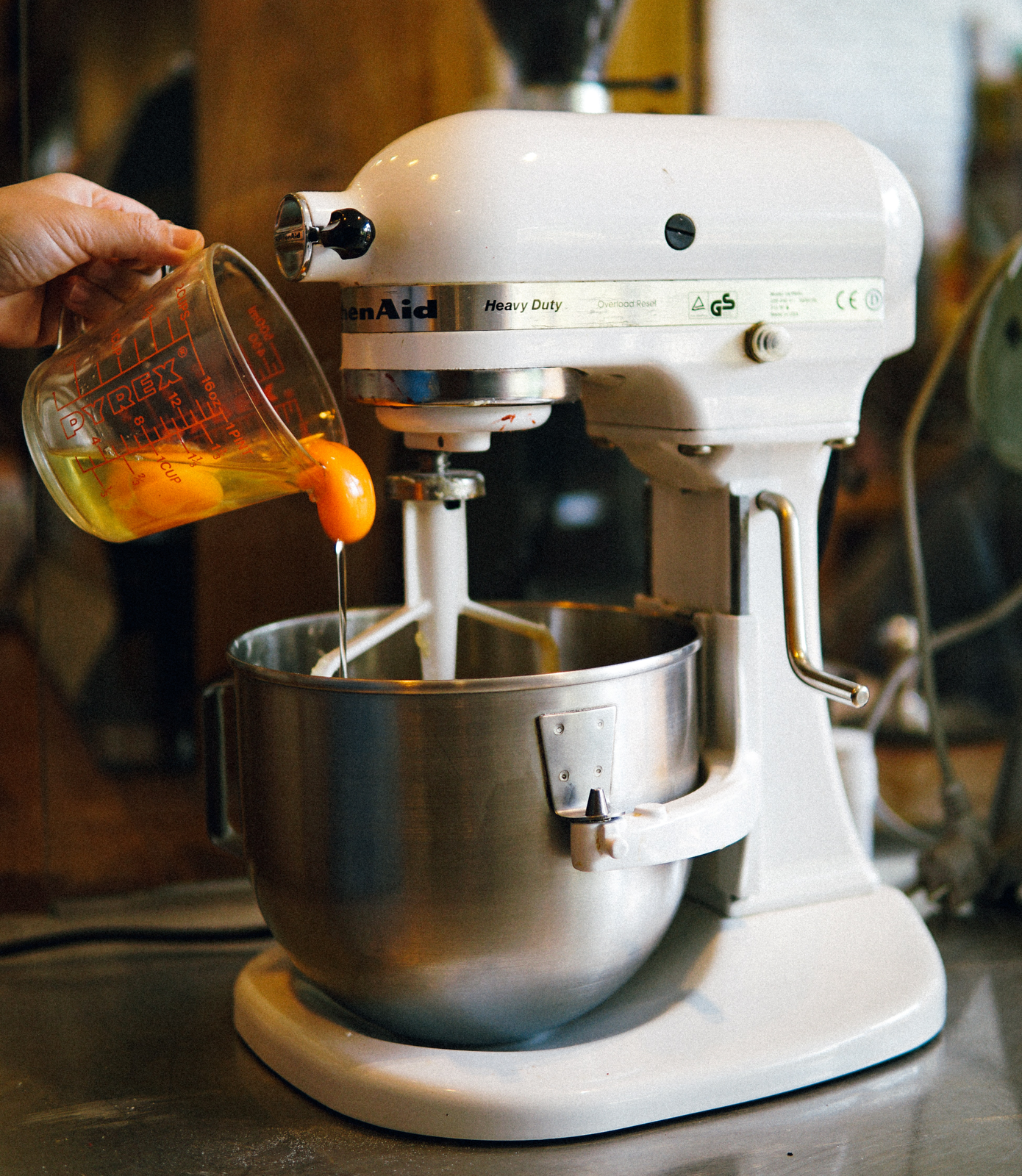Na zdjęciu widoczny jest mikser stojący. Podstawiony jest pod niego garnek. Nad garnkiem widać dłoń trzymającą naczynie z miarką. W naczyniu znajdują się dwa żółtka pływające w białku.