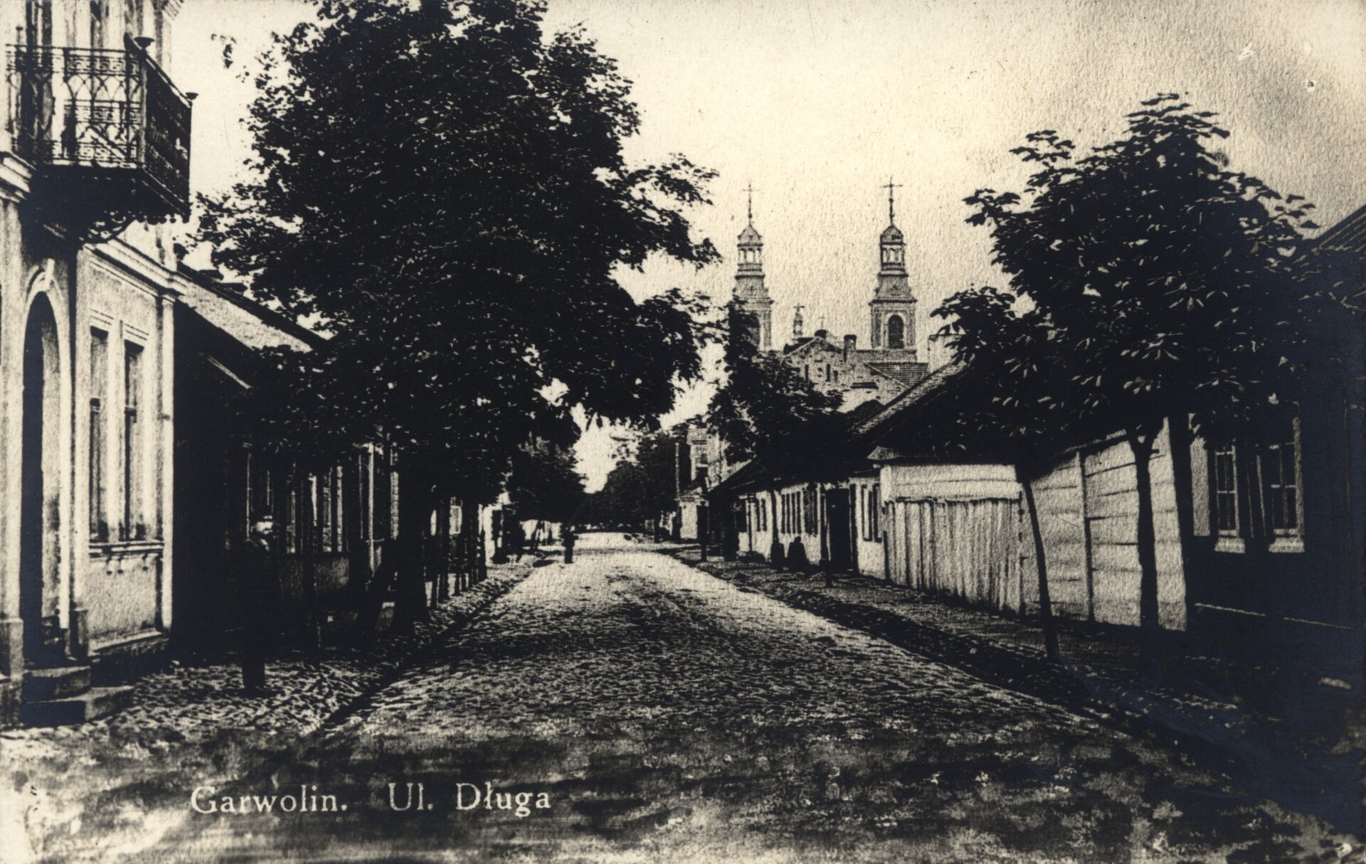 Kliknij, aby powiększyć Zdjęcie przedstawia wybrukowaną ulicę z rynsztokami i chodnikami dla pieszych, biegnącą przez jego środek. Po lewej stronie widoczny jest fragment kamienicy dwukondygnacyjnej z balkonem nad wejściem. Balustrada balkonu jest kuta. Po obu stronach ulicy są drzewa. Drewniane, parterowe domy ciągną się aż do katedry Przemienienia Pańskiego, która jest zlokalizowana po prawej stronie w głębi ulicy.