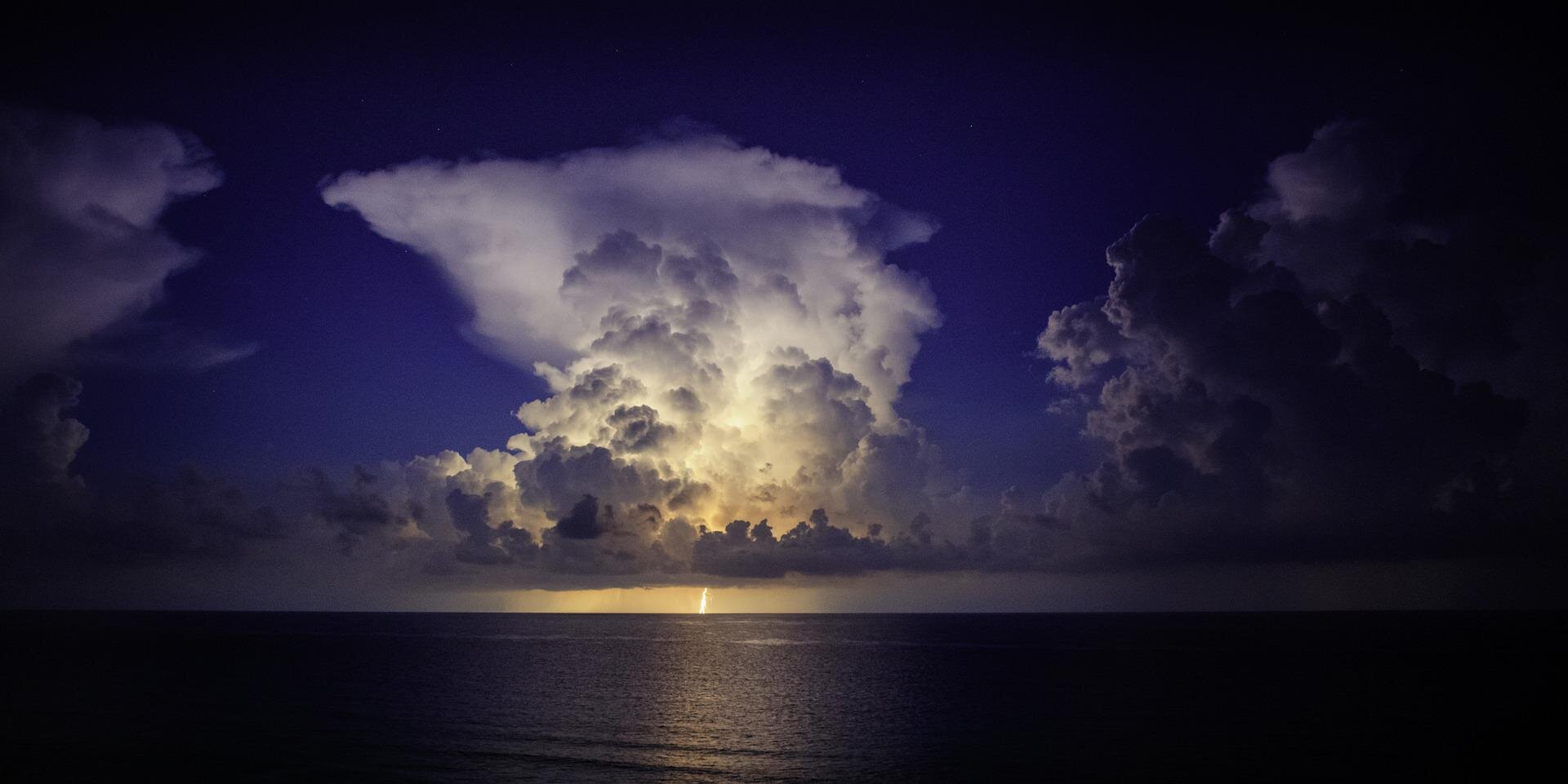 Zdjęcie przedstawia komórkę burzową. Słup chmur z rozległym kowadłem w górnej części chmury. Na pierwszym planie niemal gładka tafla morza. 