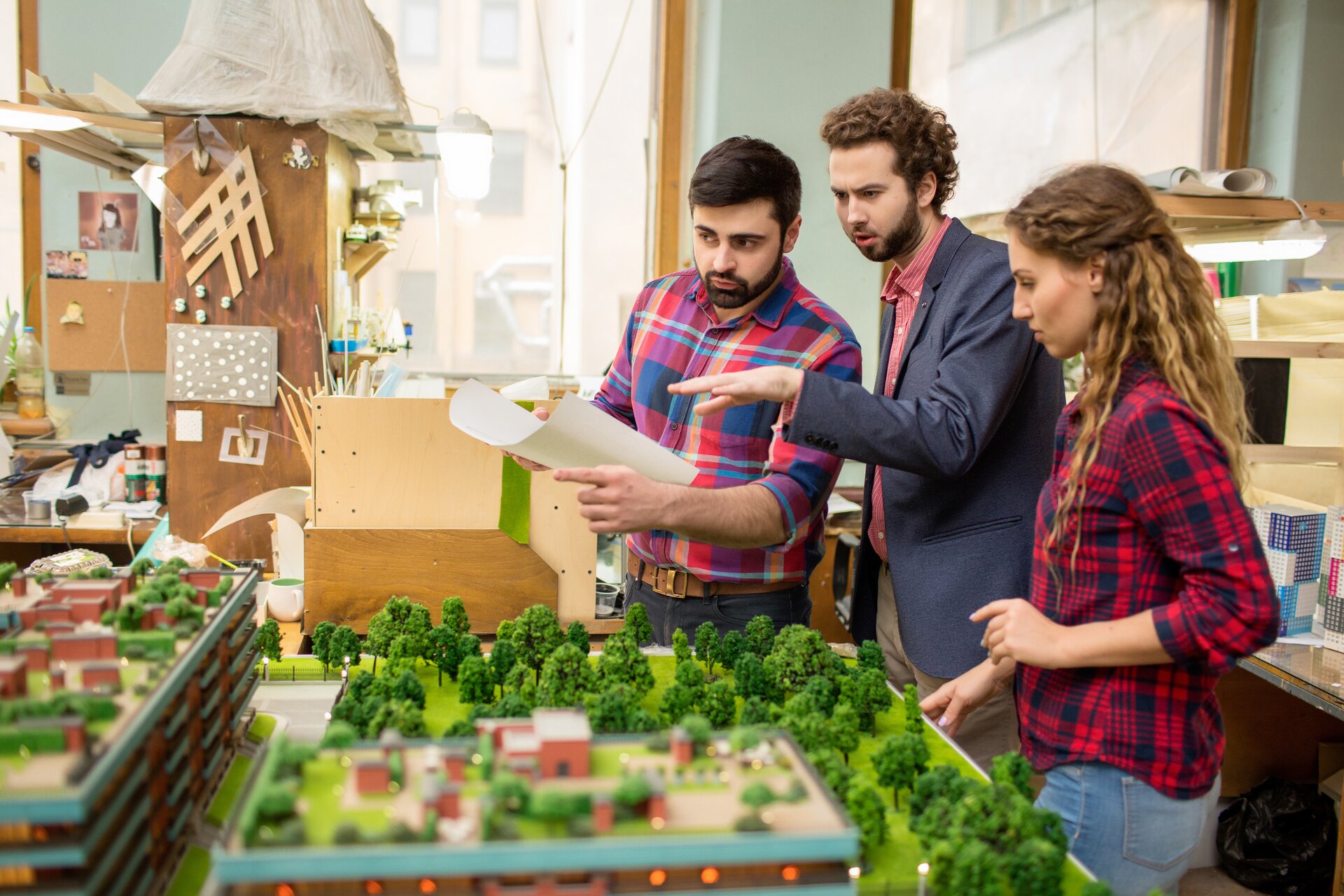 Przedstawiona fotografia ukazuje trójkę ludzi stojących przy makiecie miasta. Kobieta o długich, kręconych, blond włosach ubrana jest w czerwoną koszulę w czarną kratkę oraz niebieskie spodnie. Obok niej stoi mężczyzna o krótkich, kręconych, brązowych włosach oraz zaroście. Ubrany jest w różową koszulę, granatową marynarkę oraz brązowe spodnie. Na końcu znajduje się mężczyzna o czarnych włosach oraz zaroście, który trzyma białą kartkę. Ubrany jest w koszulę w czerwono‑granatowo‑białą kratę oraz ciemne spodnie. Makieta składa się z dwóch pięter. Pokryta jest licznymi budynkami oraz roślinnością. Pomieszczenie, w którym znajdują się ludzie zapełnione jest drewnianymi materiałami, kolorowymi papierami oraz drewnianymi półkami i szafką. Ściana pomieszczenia jest jasnozielona, pokryta drewnianymi oknami.