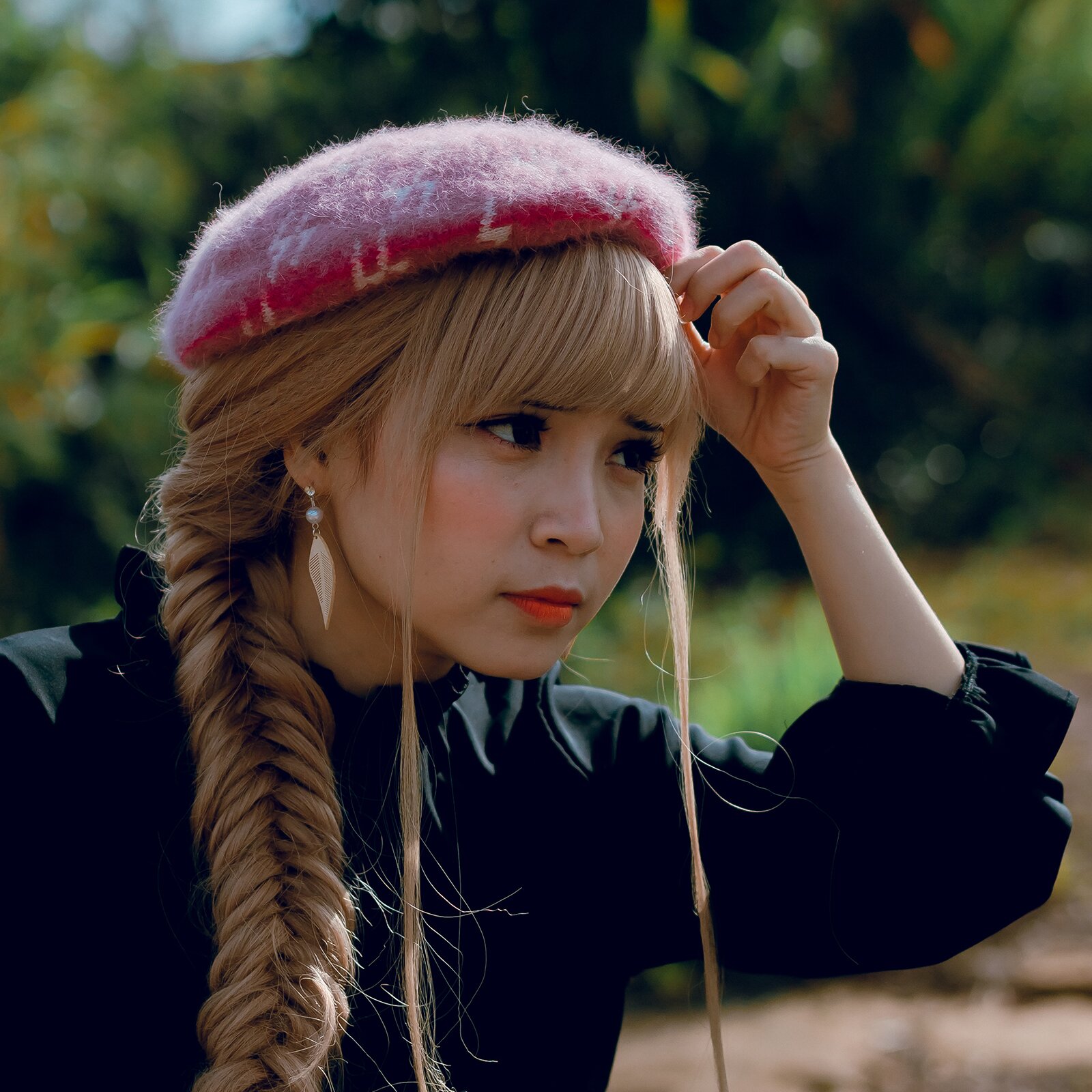 Zdjęcie przedstawia młodą kobietę o czarnych oczach z długimi rzęsami. Ma włosy blond upięte w długi warkocz. Ma grzywkę i dwa luźne pasma włosów puszczone po bokach. Jej usta pomalowane są na pomarańczowy kolor. Jest szczupła. Jest ubrana w czarną koszulę i różowy beret.