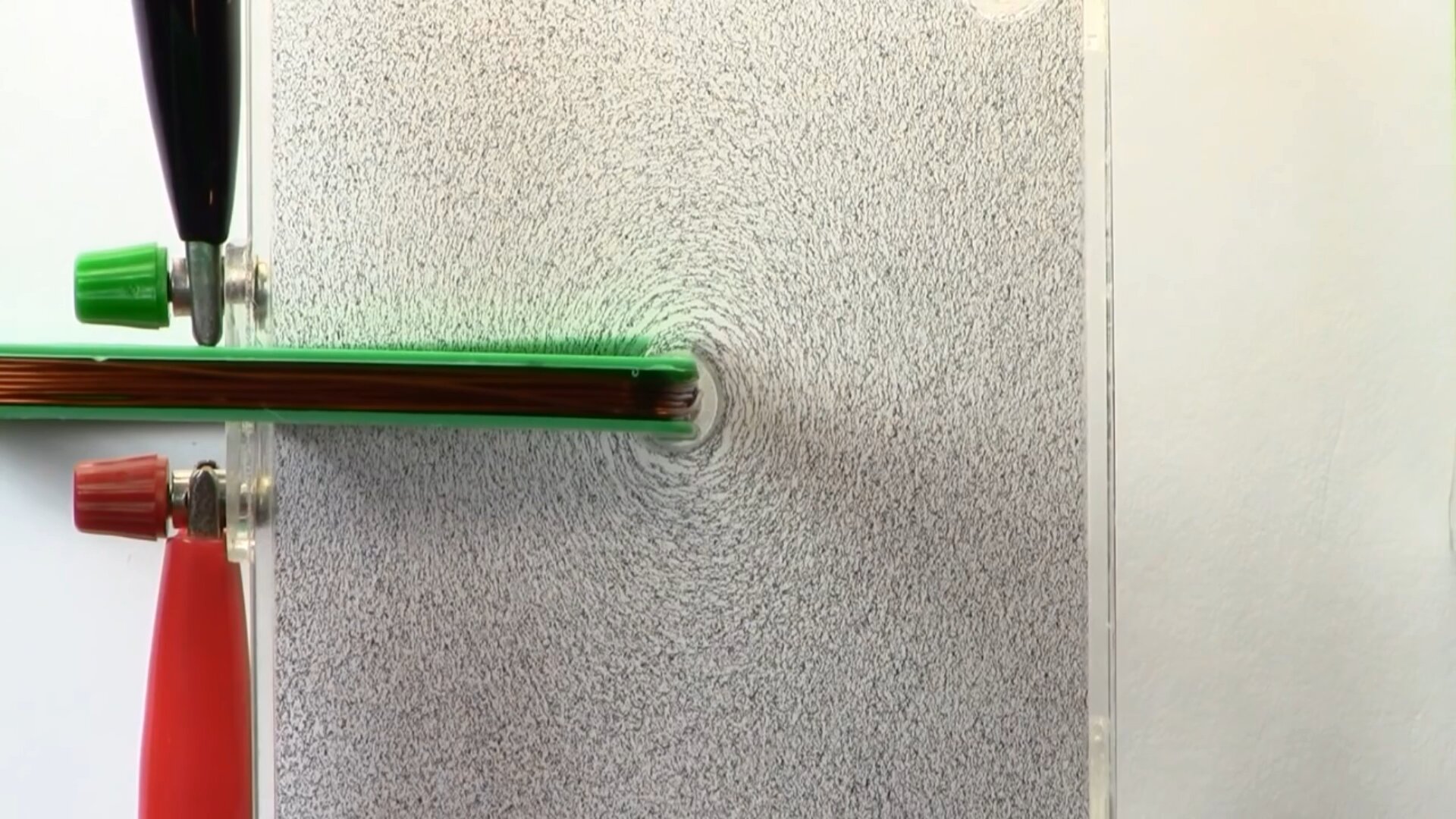 Zdjęcie przedstawia linie pola magnetycznego wokół przewodnika liniowego. Widoczna jest największa powierzchnia przezroczystej płytki, wypełnionej w środku olejem i opiłkami żelaza. Przez centralną część płytki przewleczono przewodnik tak, że jest on prostopadły do powierzchni płytki. Przewód podłączono pod prąd, dzięki czemu opiłki ułożyły się na okręgach wokół fragmentu przewodnika przechodzącego przez płytkę.