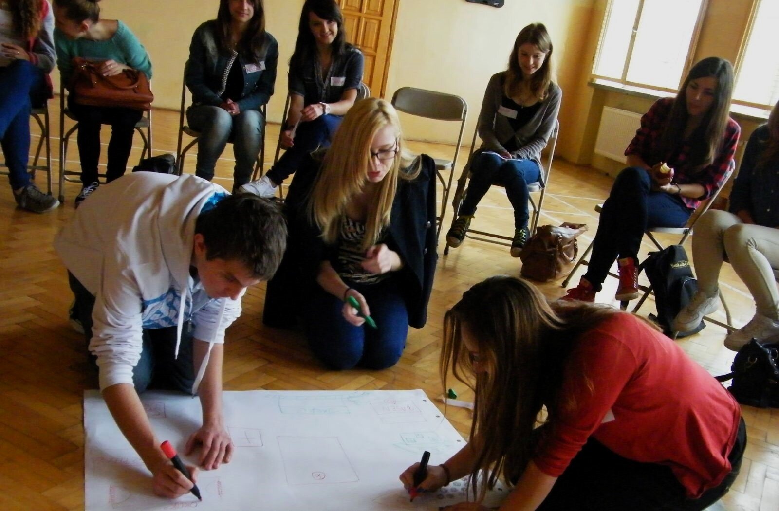 Zdjęcie pokazuje młodzież podczas warsztatów. Na krzesłach ustawionych w okręgu siedzą młodzi ludzie, rozważają pomysły, dyskutują. Wewnątrz okręgu, kilka osób zapisuje wszystko na dużym kartonie, leżącym na podłodze.