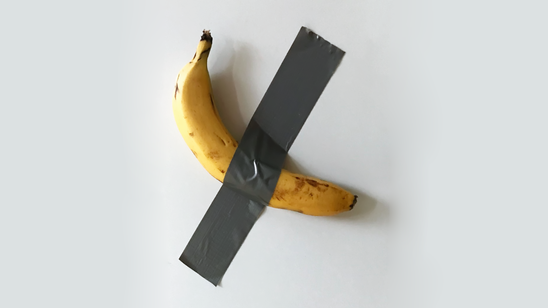 Zdjęcie przedstawia dzieło Maurizia Cattelana. Jest to banan przyklejony do ściany paskiem taśmy klejącej.