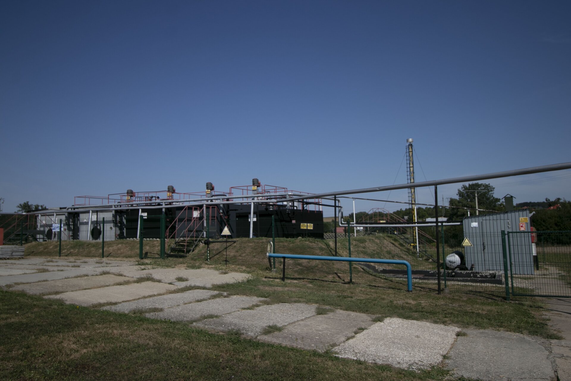 Zdjęcie przedstawia zespół prostopadłościennych zbiorników na ropę naftową wraz z orurowaniem. Na pierwszym planie chodnik. W tle błękitne niebo.