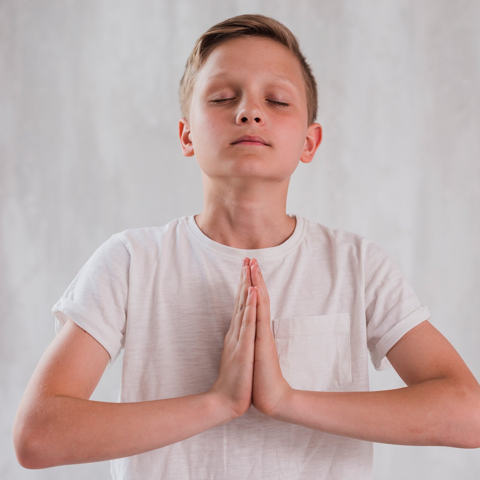 Ilustracja przedstawia chłopca w wieku szkolnym, który ubrany jest w jasną koszulkę z krótkim rękawem. Chłopiec ma zamknięte oczy i dłonie złożone razem jak do modlitwy.  Skupia się na oddechu.
