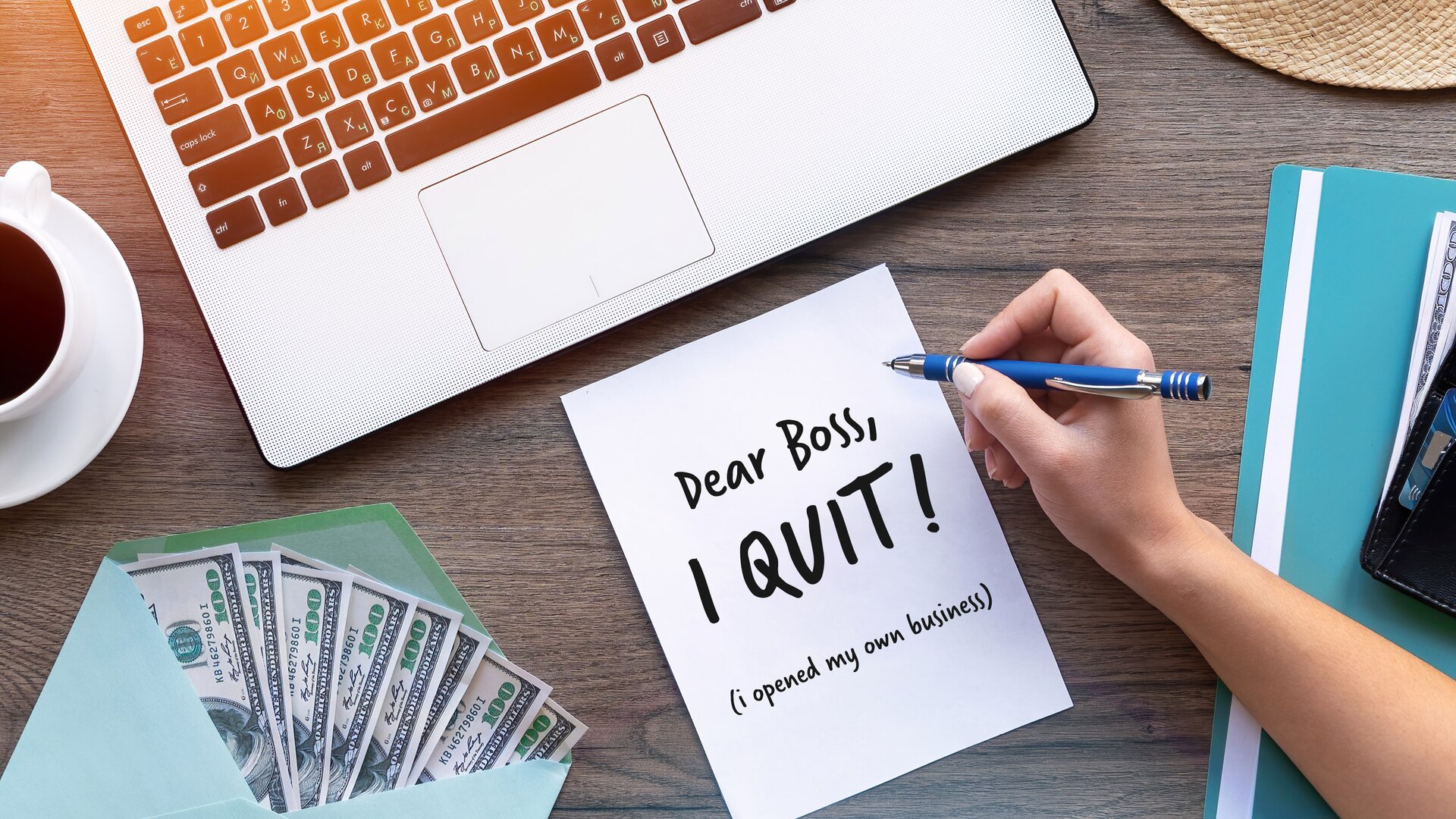 Zdjęcie przedstawia blat, na którym ustawiono laptop, a obok niego filiżankę z ciemnym napojem.   Przed komputerem leży karta z napisem "Dear Boss, I QUIT! (I opened my own business)".   Nad kartką widoczna jest ręka trzymająca długopis.   Obok leży koperta, z której wystają studolarowe banknoty.   Zdjęcie zostało wykonane z góry, co pozwala dobrze uchwycić wszystkie szczegóły kompozycji.