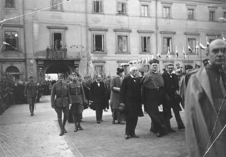 Fotografia przedstawia grupę mężczyzn zmierzających w jednym kierunku. Są wśród nich biskupi, wojskowi oraz cywile ubrani w eleganckie stroje, garnitury, koszule i krawaty. Niektórzy rozmawiają ze sobą, inni idą w skupieniu. Wokół widać żołnierzy ustawionych w równych rzędach. Z tyłu widać budynek z oknami i balkonem gdzie widać przyglądających się całej scenie ludzi.