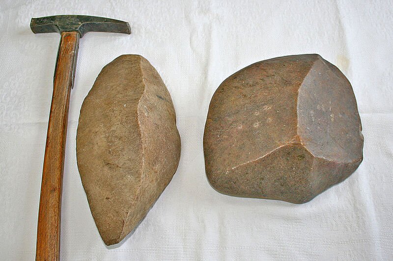 Na zdjęciu są dwa kawałki odłamków skalnych o wygładzonych ścianach i dwóch oraz trzech ostrych krawędziach. Obok skal leży młotek. 