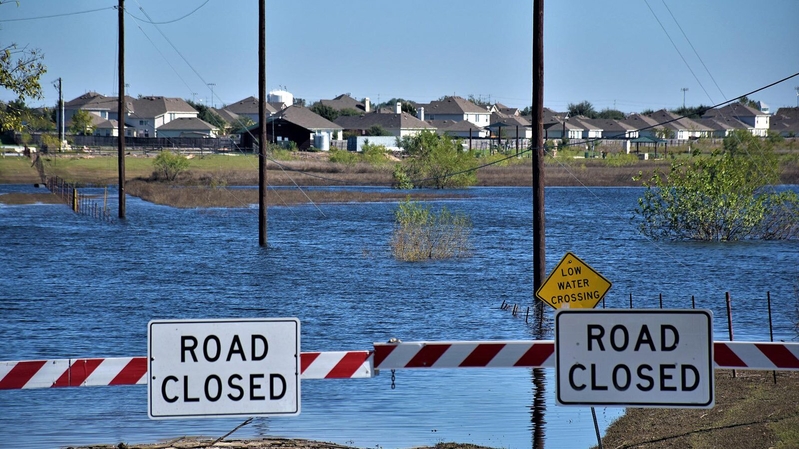 Zdjęcie przedstawia biało‑czerwone poziome belki, na których umieszczono tabliczki z napisem "ROAD CLOSED", a za nimi stoi żółty znak na którym napisano "LOW WATER CROSSING". Za znakami ukazaną dużą przestrzeń zalaną przez wodę, z której wystają fragmenty krzewów oraz słupów. W oddali są domy jednorodzinne.