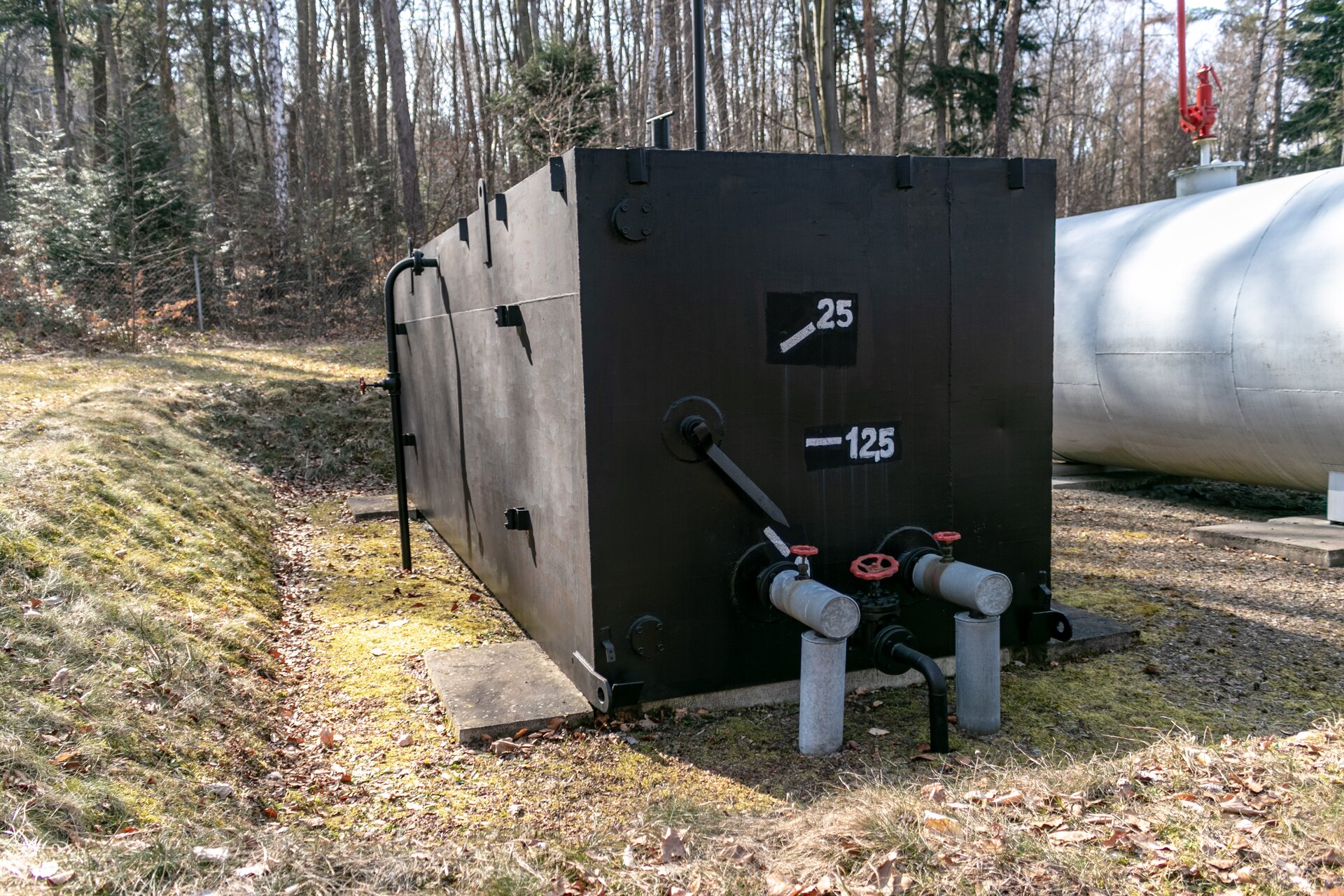 Na zdjęciu widać zbiornik napowierzchniowy skrzyniowy o pojemności dwudziestu pięciu metrów sześciennych jest do magazynowania ropy naftowej lub wody złożowej. Zbiornik wykonany jest ze stali w kolorze czarnym. Na froncie zbiornika w miejscu odpowiadającym połowie jego wysokości oraz w miejscu odpowiadającym około siedemdziesięciu procentom jego wysokości znajdują się białe liczby dwanaście przecinek pięć oraz dwadzieścia pięć. Do frontu zbiornika przyłączone są dwie stalowe rury. Pomiędzy nimi biegnie dodatkowa czarna rura o mniejszej średnicy. Wszystkie rury wyposażone są u góry w czerwone kurki służące do regulacji przepływu. Za zbiornikiem skrzyniowym widać fragment stalowego zbiornika cylindrycznego. W tle zbiornia znajduje się roślinność leśna.