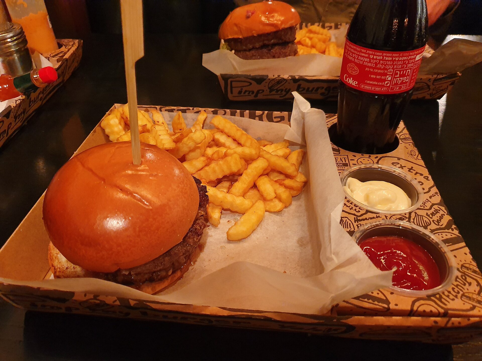 Zdjęcie przestawia tacę, na której leży duży hamburger, w który jest wbita drewniana wykałaczka, karbowane frytki, dwa sosy: biały i czerwony i butelka z Cola‑Colą. W tle widać dwie inne tace z podobnymi zestawami.