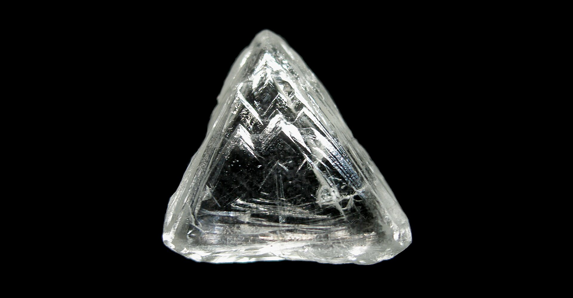 Zdjęcie przedstawia idealnie bezbarwny, nieoszlifowany diament w kształcie nieregularnego stożka na czarnym tle.