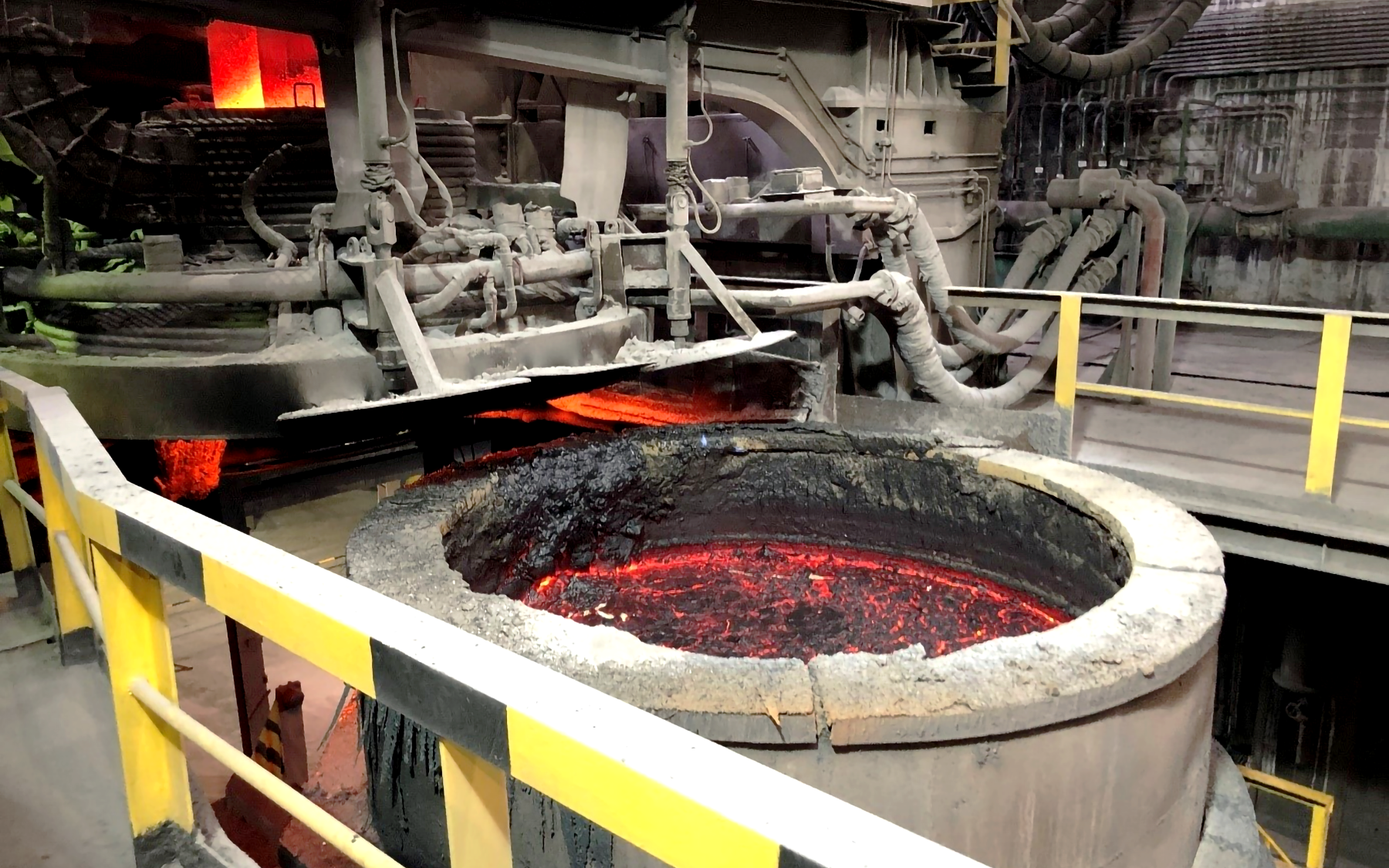 Obrazek przedstawia fotografię piecokadzi, czyli stanowiska służącego do rafinacji elektrodami grafitowymi. Widoczna jest kadź o walcowatym kształcie, wypełniona ciekłym metalem, na którego powierzchni widać ciemny kożuch tlenków. Kadź nie jest wielka, ma około metra wysokości i mniej niż metr średnicy. Nad nią i za jej obręczą umieszczone są elementy maszynerii rafinującej, składająca się z przewodów, rur i profili.