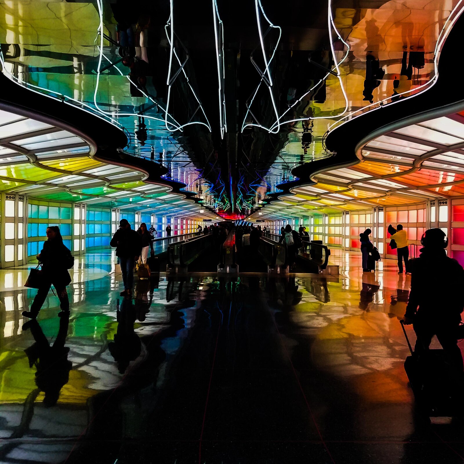 Zdjęcie przedstawia tunel prowadzący pasażerów na lotnisko ukazany w perspektywie zbieżnej. Jest cały oszklony, ściany  boczne zbudowane są z tafli kolorowego szkła, które odbija się w podłodze. Sufit składa się z przezroczystych płyt o nieregularnej formie, które również odbijają różnobarwne światło przechodzące przez kolorowe ściany. Pośrodku sufitu delikatnie świecą wijące się wężyki dodatkowych lamp. Tunelem podążają pasażerowie z bagażami, w tym oświetleniu można zauważyć tylko czarne sylwetki. 